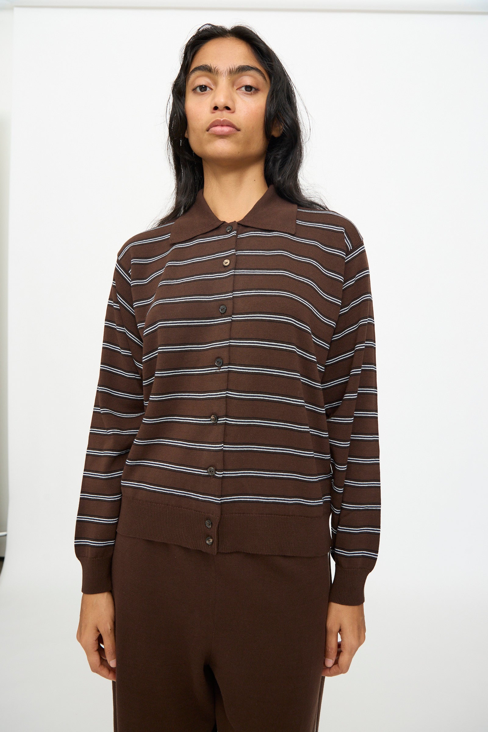 A person is wearing the Cordera Cotton Polo Cardigan in Striped—a brown, long-sleeve cardigan crafted from soft organic cotton with horizontal white stripes—paired with matching brown pants, standing against a plain white background.
