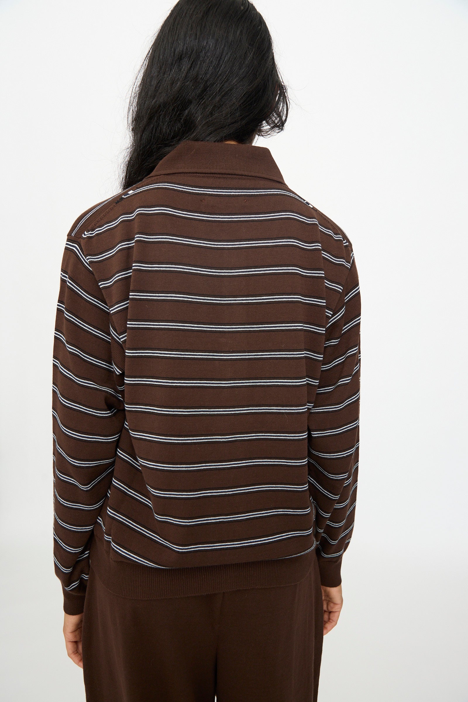 A person stands with their back to the camera, wearing the Cordera Cotton Polo Cardigan in Striped—a brown organic cotton long-sleeve shirt with white horizontal stripes—paired with matching brown pants against a plain background.