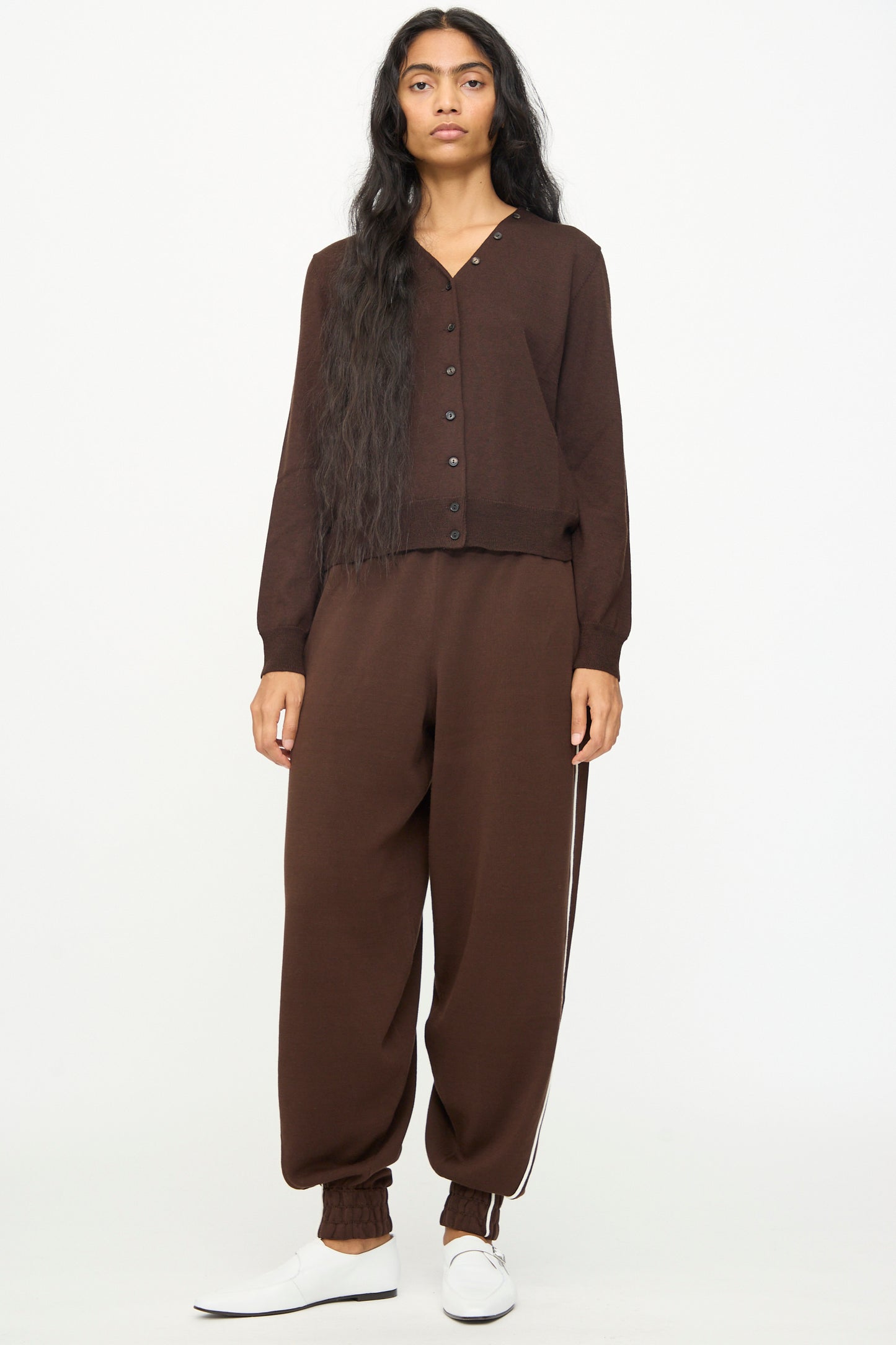 A person with long dark hair models Cordera's Cotton Sporty Pants in Brown, featuring an elastic waistband and white stripe, paired with a matching brown cardigan and white slip-on shoes against a white background.