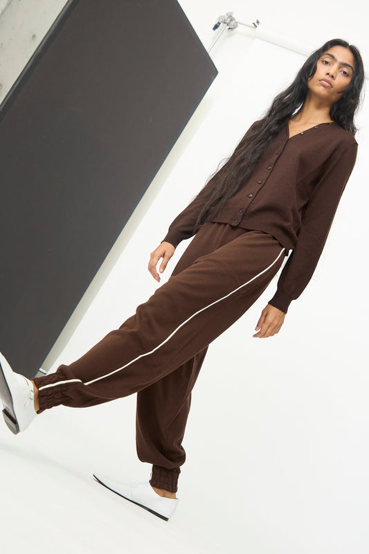 A woman models Cordera’s Cotton Sporty Pants in Brown, styled with a coordinating buttoned cardigan and white shoes, against a white background. The knit pants feature white side stripes for a sporty touch.