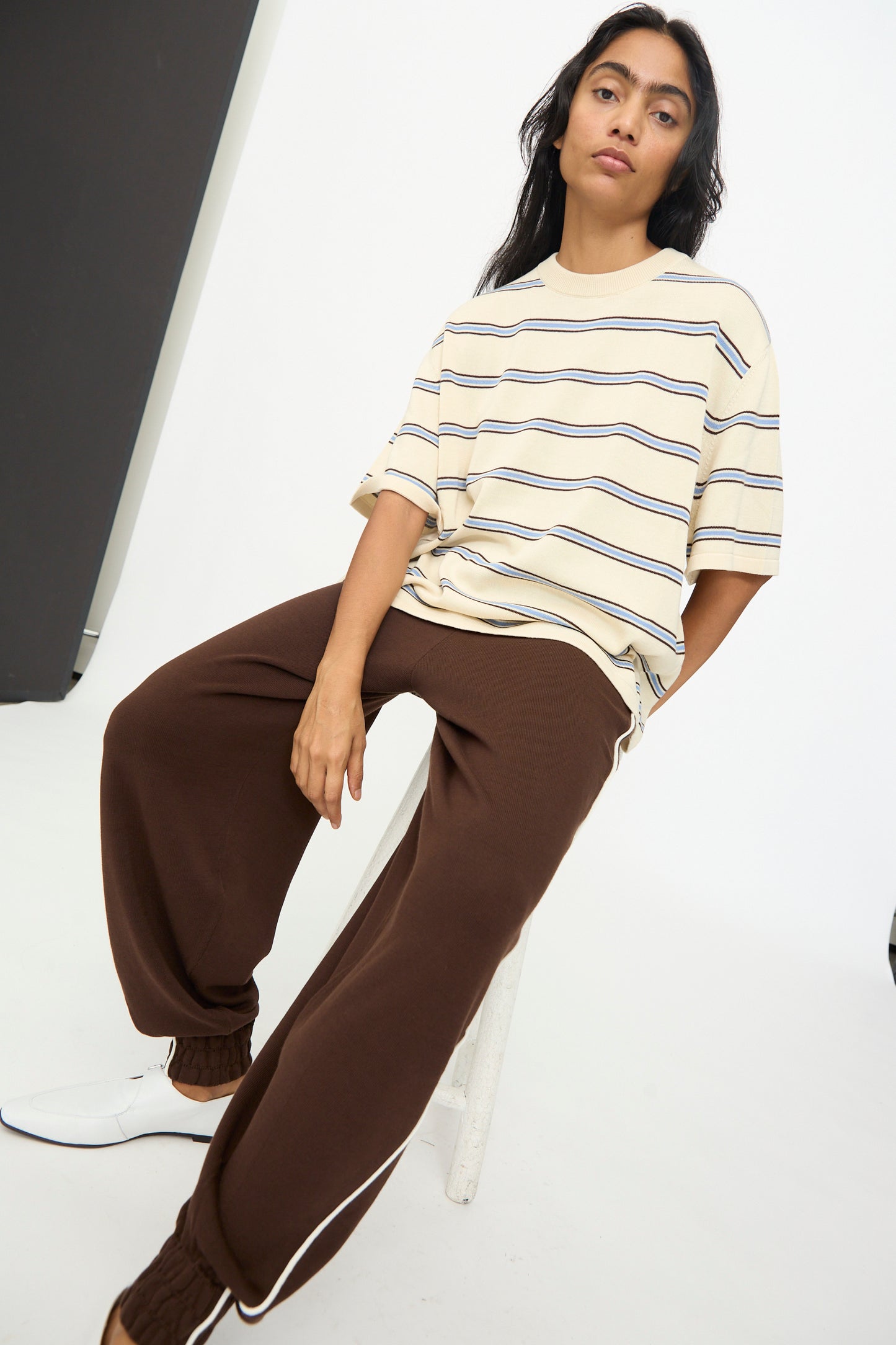 A person seated on a white stool wears the Cordera Cotton T-Shirt in Striped—an oversized, beige cotton design made in Spain—paired with brown sweatpants and white shoes, all set in a minimalist studio environment.
