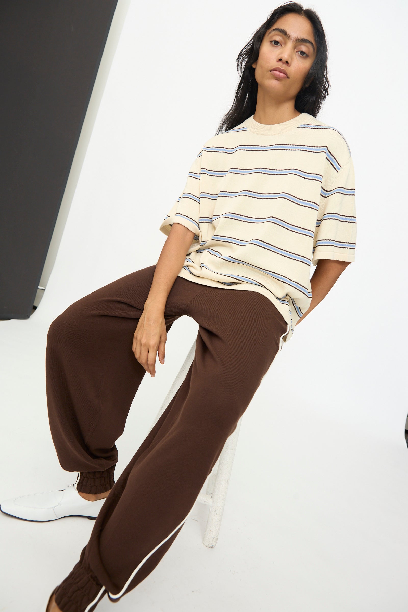 A person seated on a white stool wears the Cordera Cotton T-Shirt in Striped—an oversized, beige cotton design made in Spain—paired with brown sweatpants and white shoes, all set in a minimalist studio environment.