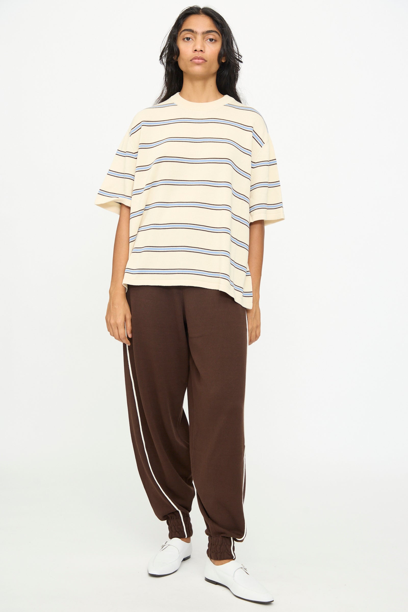 A person stands against a white background, wearing the Cordera Cotton T-Shirt in Striped with an oversized fit in beige short sleeves, paired with brown track pants featuring side stripes and white shoes.