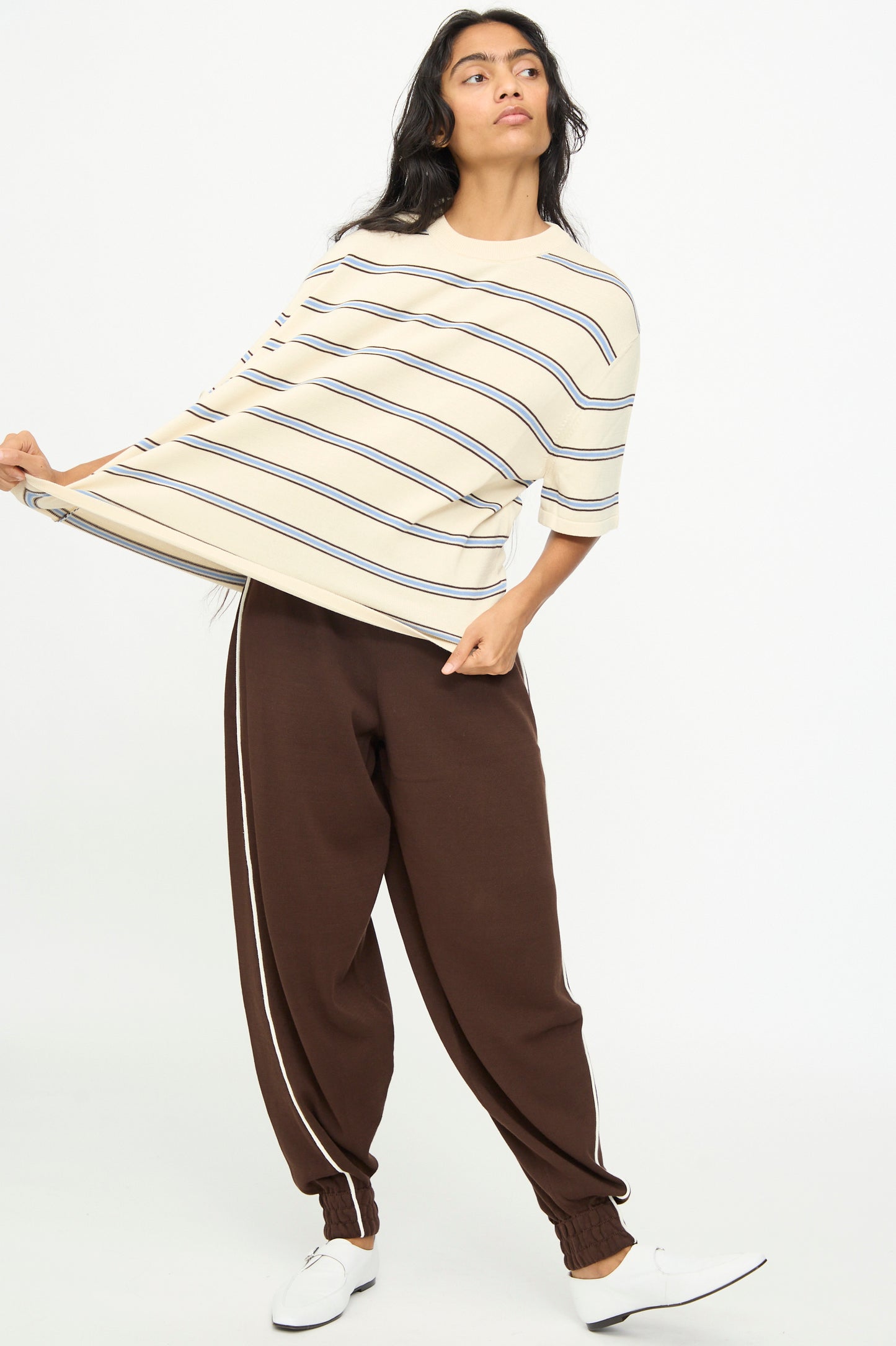 A person wearing the Cordera Cotton T-Shirt in Striped (oversized fit, cream), paired with brown jogger pants featuring white side stripes and white shoes, stands while holding out one side of the shirt.