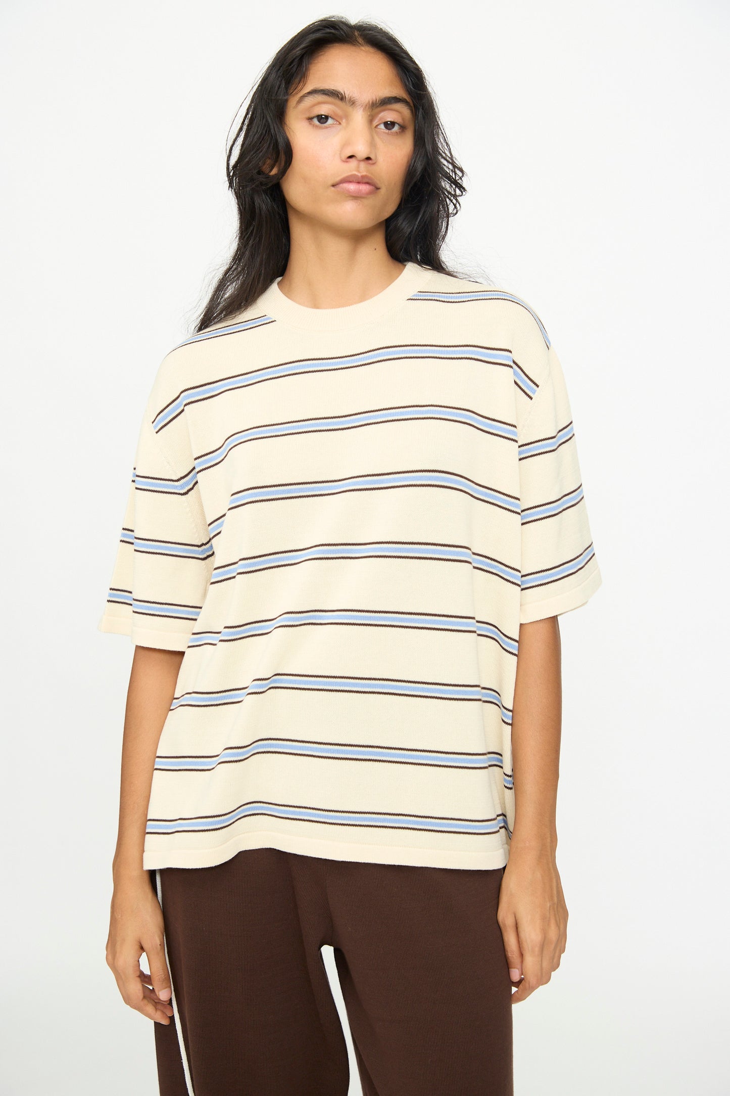 A person wearing Cordera's Cotton T-Shirt in Striped—an oversized beige short-sleeved cotton tee with dark horizontal stripes—paired with brown pants, standing against a plain white background.