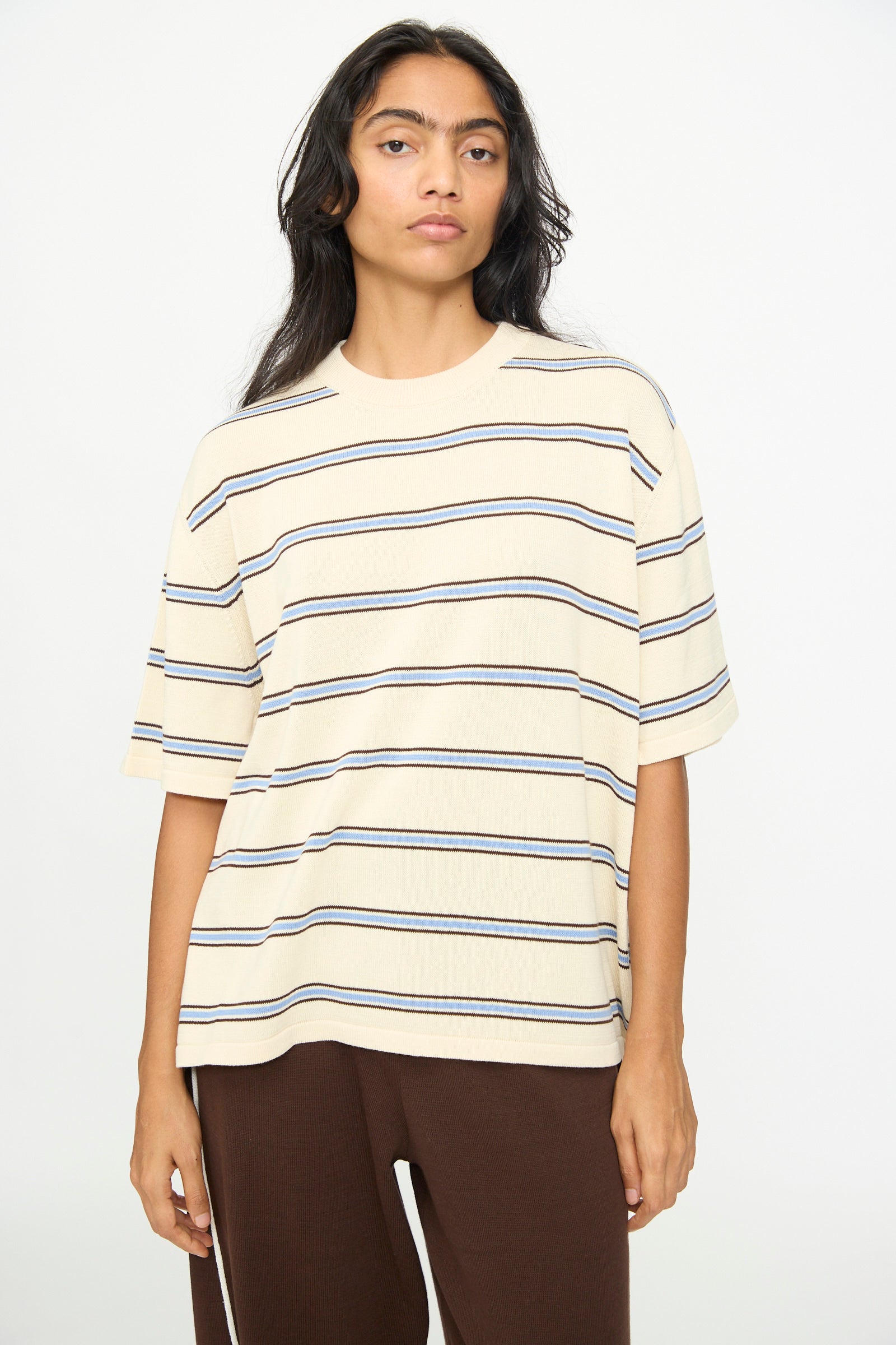 A person wearing Cordera's Cotton T-Shirt in Striped—an oversized beige short-sleeved cotton tee with dark horizontal stripes—paired with brown pants, standing against a plain white background.