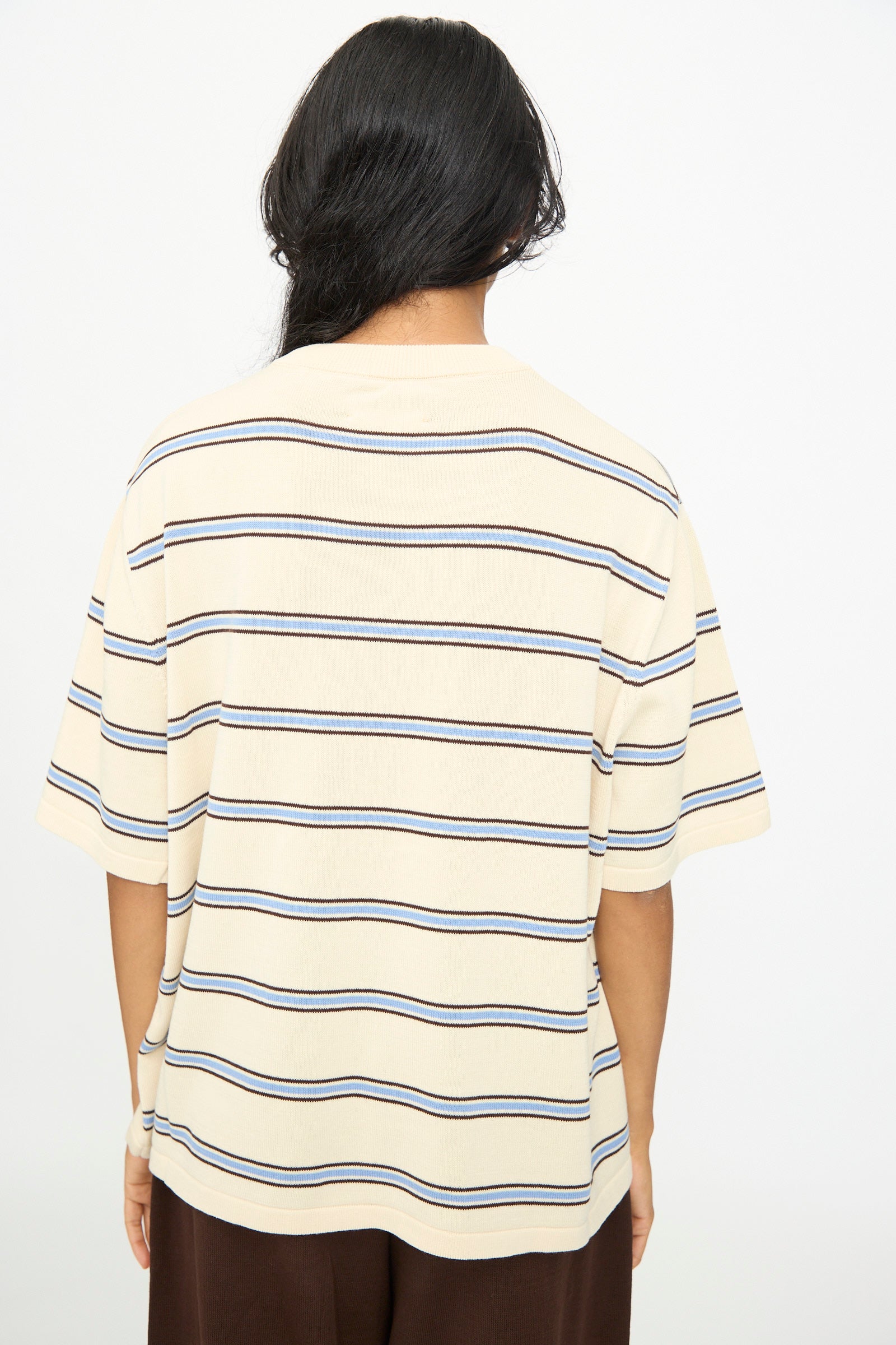 A person with long dark hair stands facing away, wearing the Cordera Cotton T-Shirt in Striped—an oversized fit beige short-sleeve cotton shirt with horizontal navy and brown stripes—paired with dark pants, set against a plain white background.