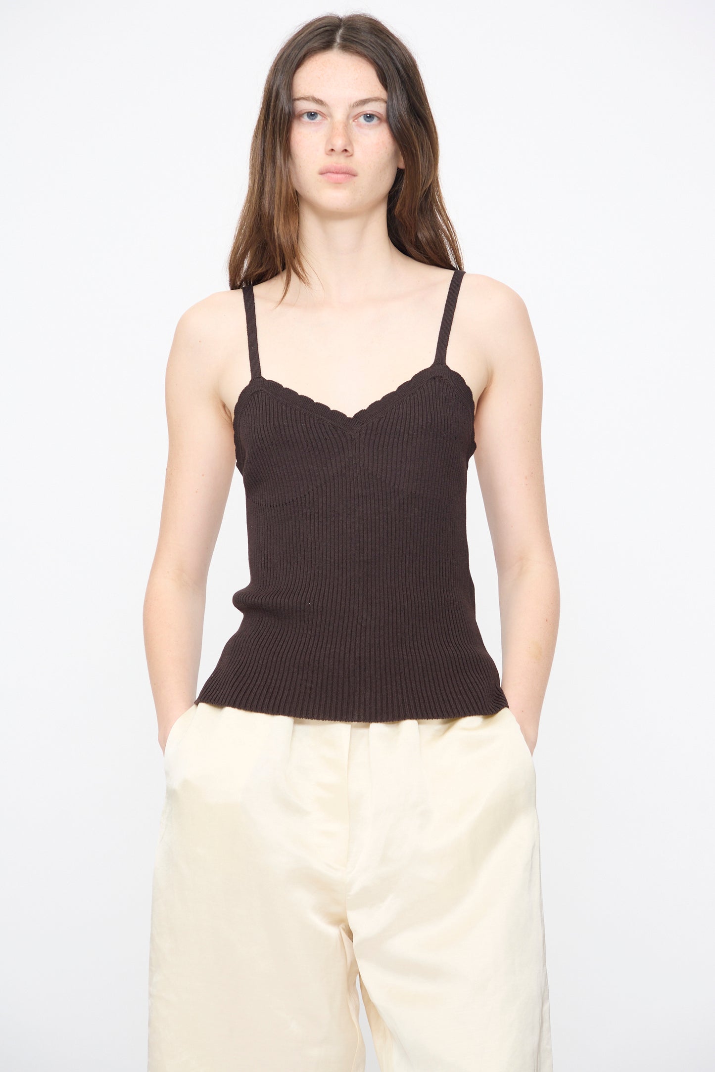 A woman with long brown hair models the Cordera Cotton and Silk Ribbed Tank Top in Brown with loose cream-colored pants, standing against a plain white background with her hands in her pockets.