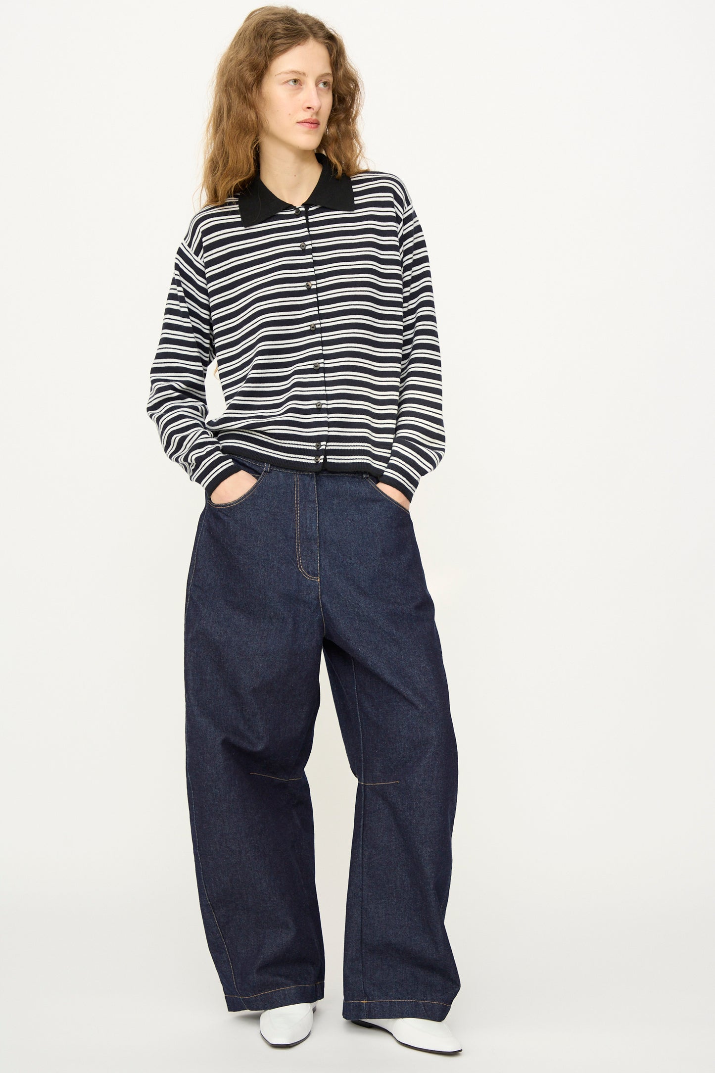 A person wearing Cordera’s Curved Baggy Jean in Denim with a black and white striped long-sleeve shirt and white shoes stands against a plain light background.