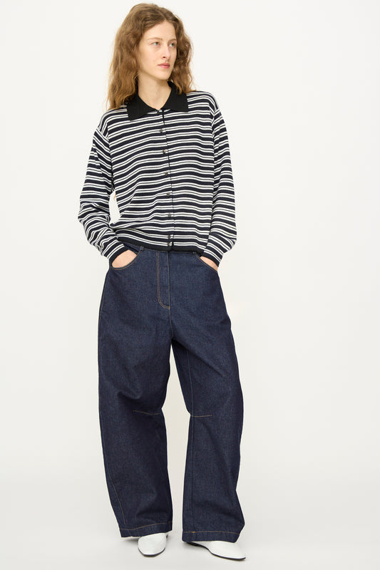 A person wearing Cordera’s Curved Baggy Jean in Denim with a black and white striped long-sleeve shirt and white shoes stands against a plain light background.