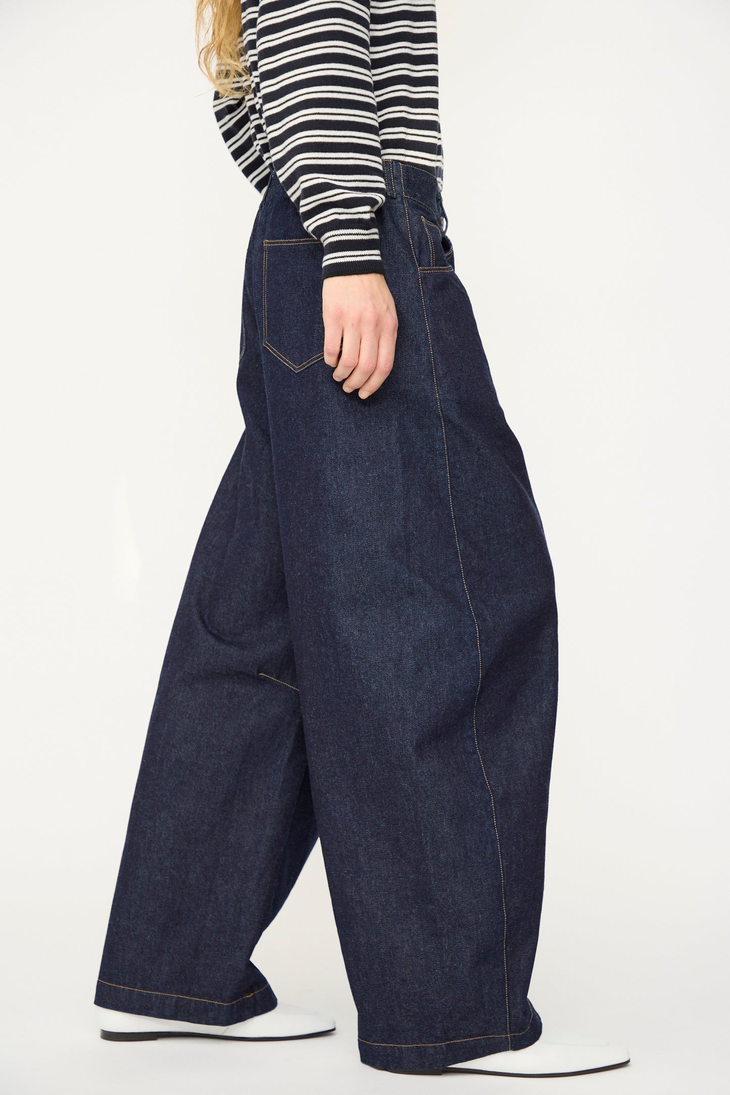 A person stands against a white background wearing Cordera's Curved Baggy Jean in dark blue organic cotton denim and a black-and-white striped long-sleeve shirt.