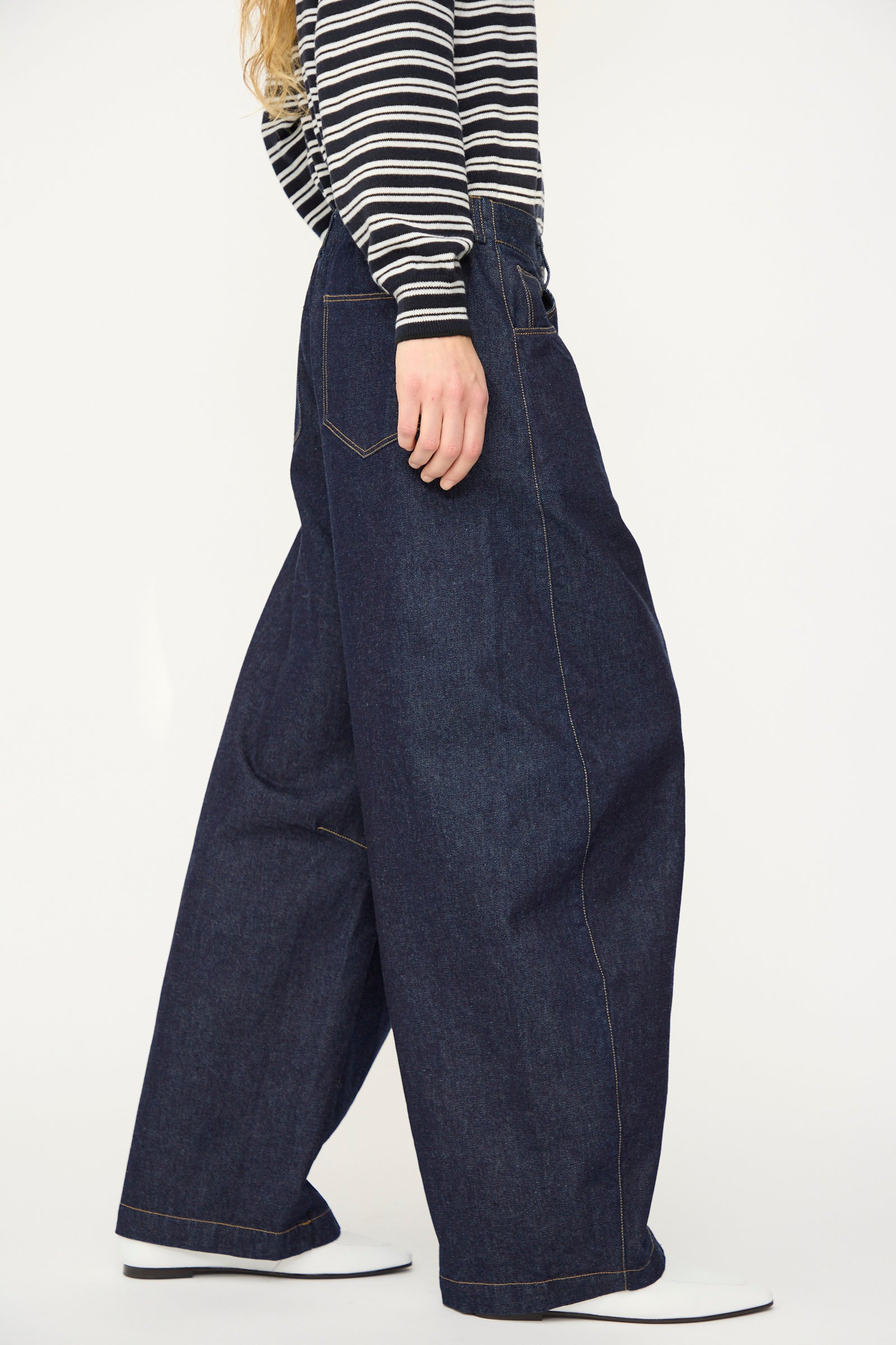 A person stands against a white background wearing Cordera's Curved Baggy Jean in dark blue organic cotton denim and a black-and-white striped long-sleeve shirt.