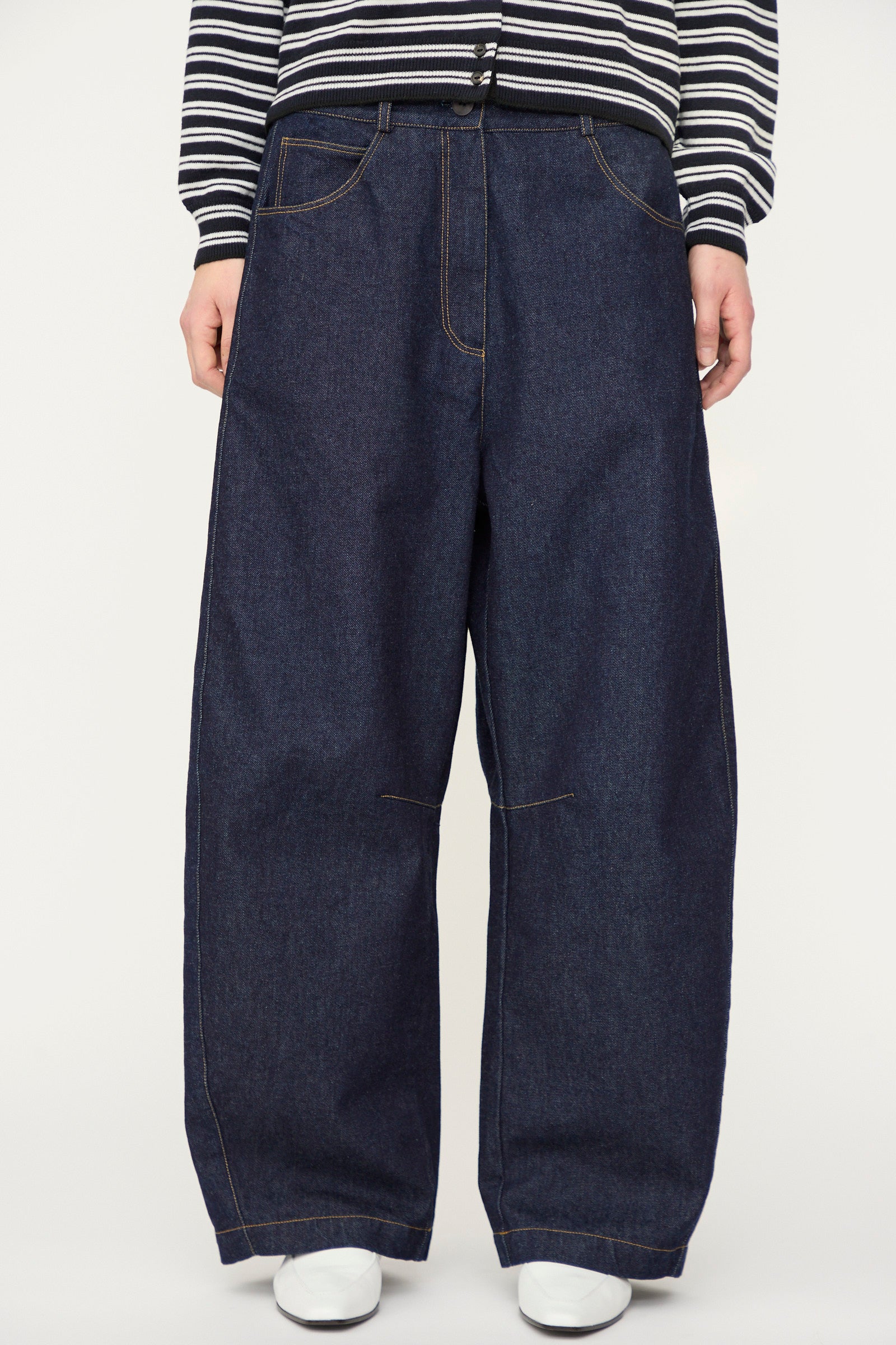A person wears Cordera's Curved Baggy Jean in dark blue denim, paired with a black-and-white striped long-sleeve top and white shoes. The photo is cropped from the shoulders down.