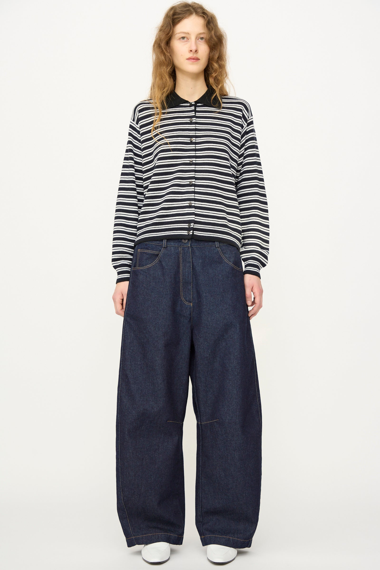 A person stands facing forward in a black and white striped long-sleeve shirt, Cordera Curved Baggy Jean in Denim, and white shoes against a plain white background.