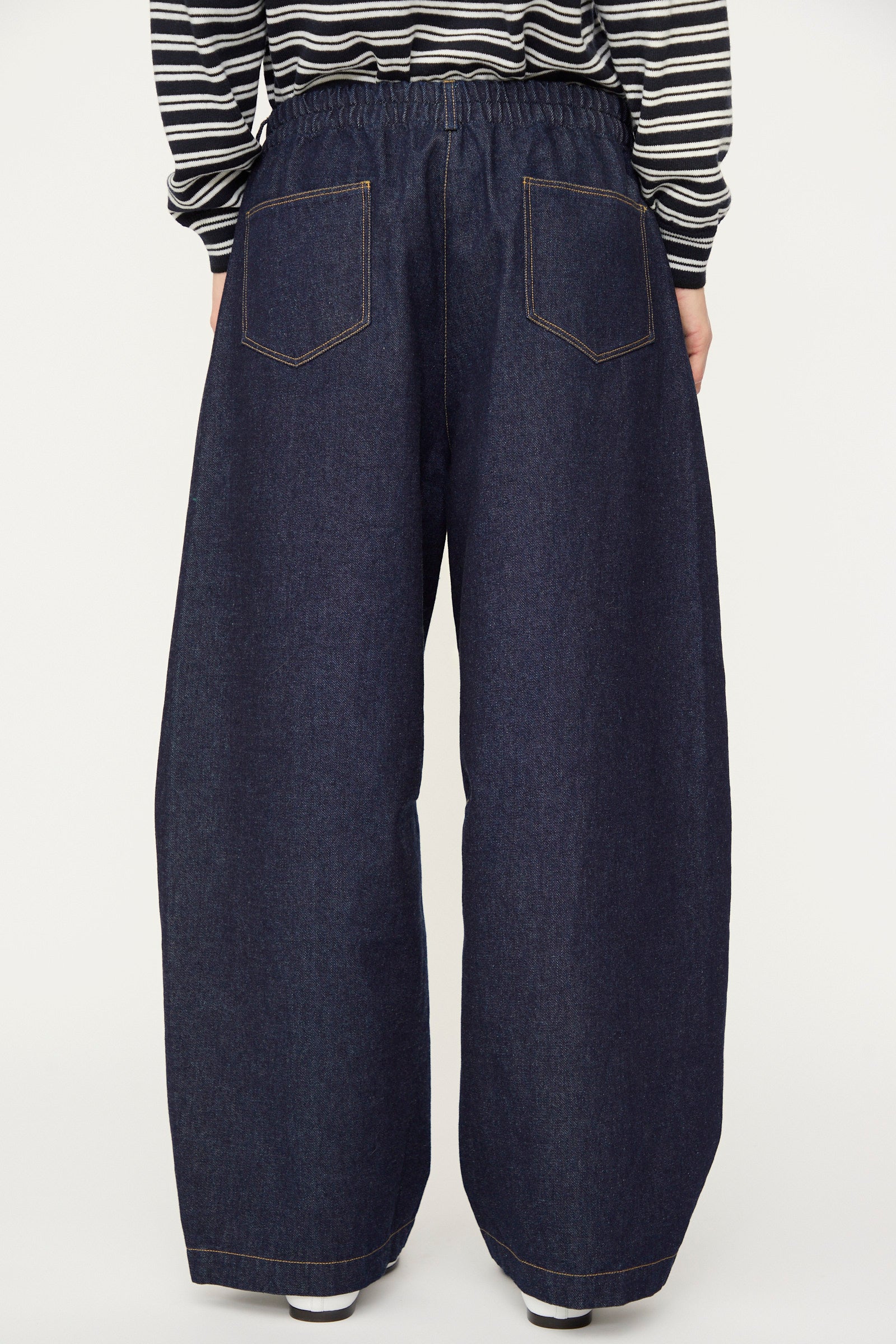 A person is shown from the back wearing Cordera’s Curved Baggy Jean in dark denim and a black-and-white striped long-sleeve shirt. The high-rise, wide-leg jeans are crafted from organic cotton.