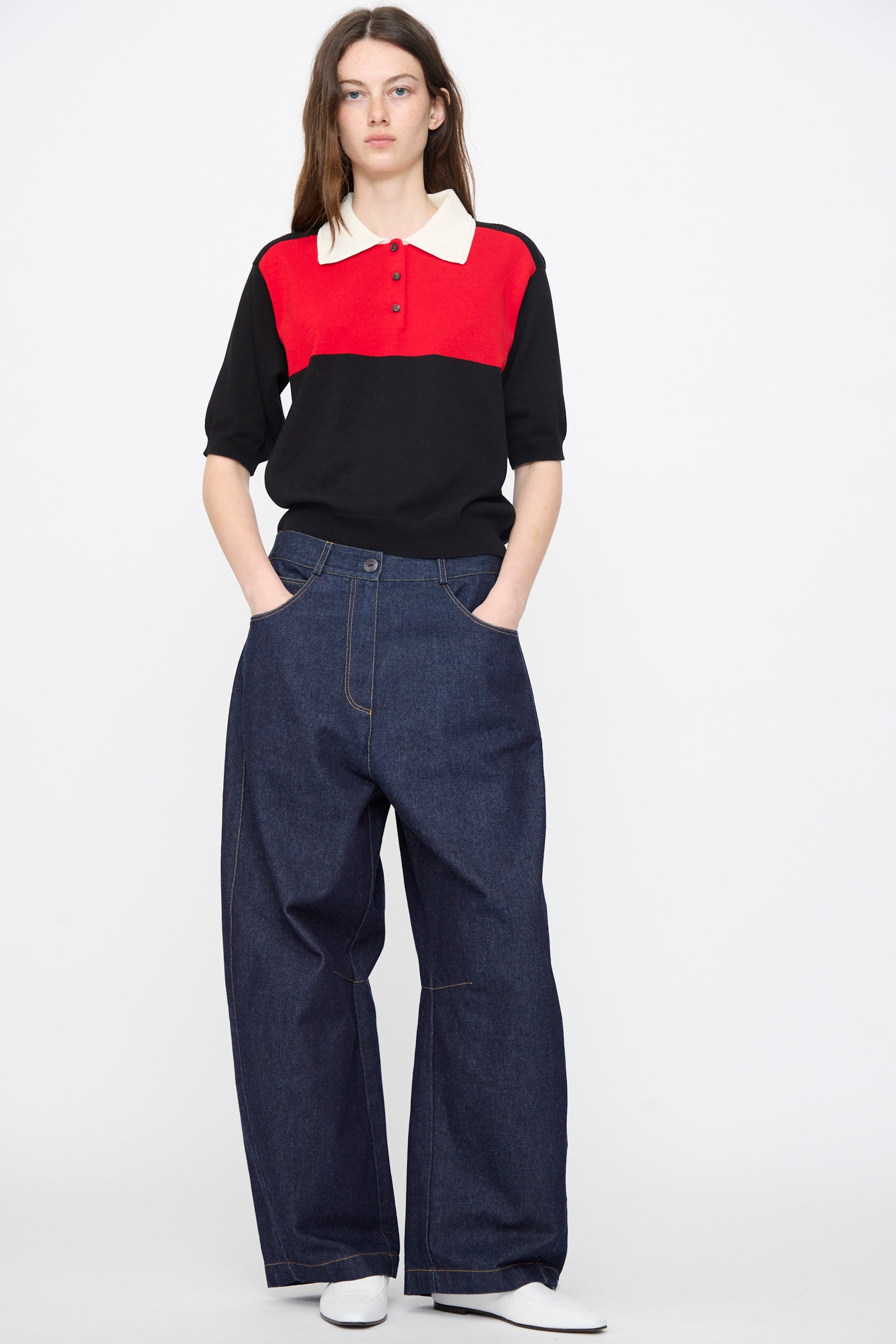 A person stands with hands in pockets, wearing Cordera's Curved Baggy Jeans in Denim and a black and red collared shirt, paired with white shoes against a plain white background.