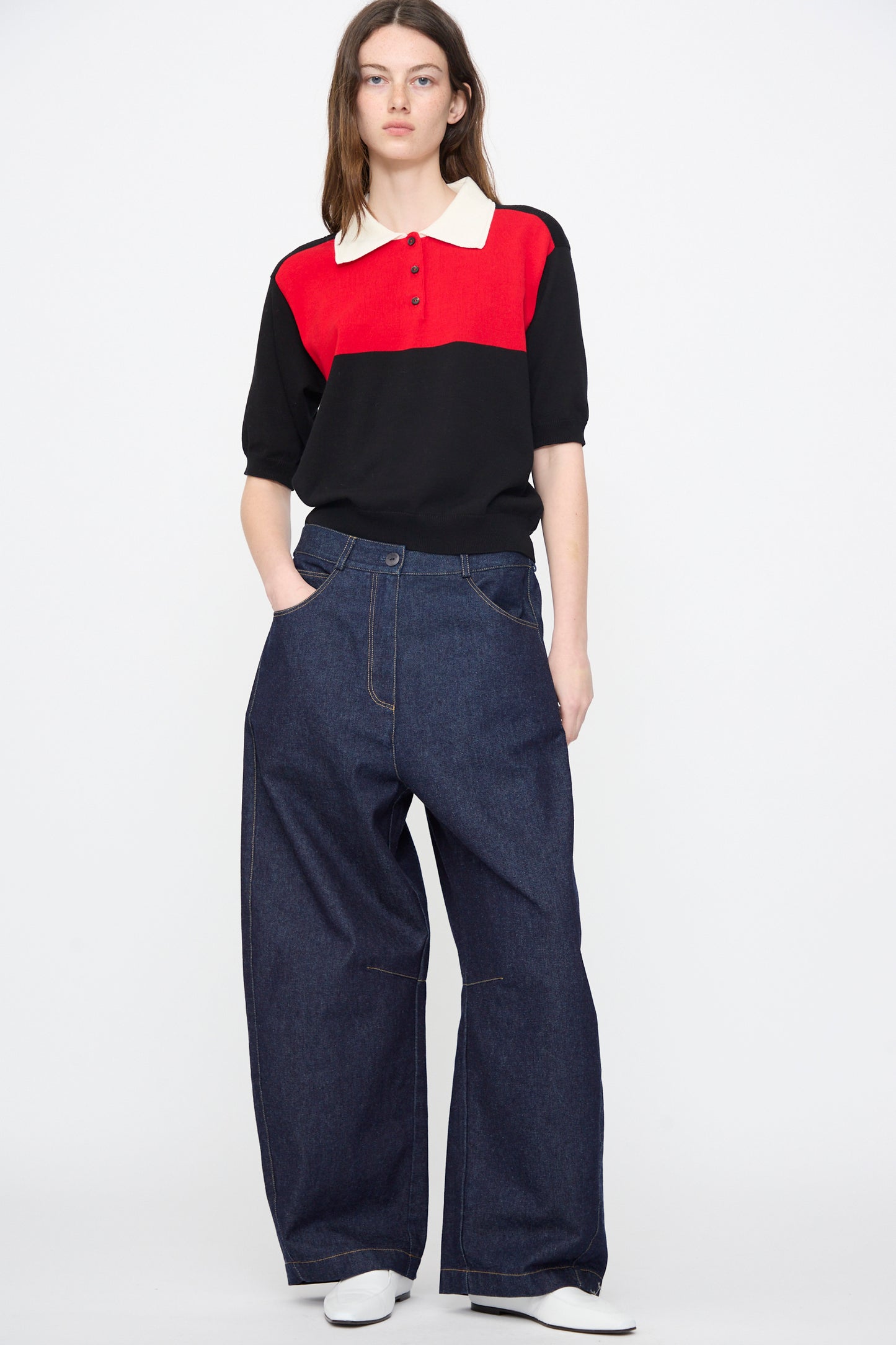 A person stands against a plain background wearing a red and black collared shirt, Cordera Curved Baggy Jeans in Denim, and white shoes, hands in pockets.