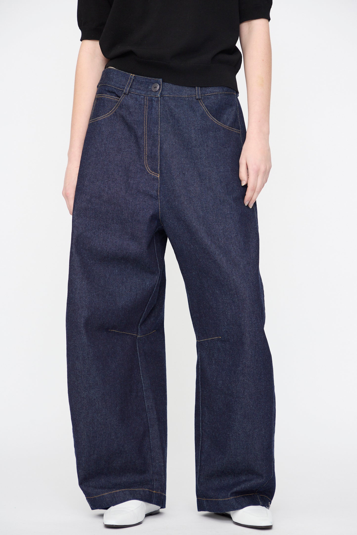Person wearing Cordera’s Curved Baggy Jeans in Denim, featuring dark blue organic cotton and a relaxed wide-leg fit, paired with a black top and white shoes against a plain white background.