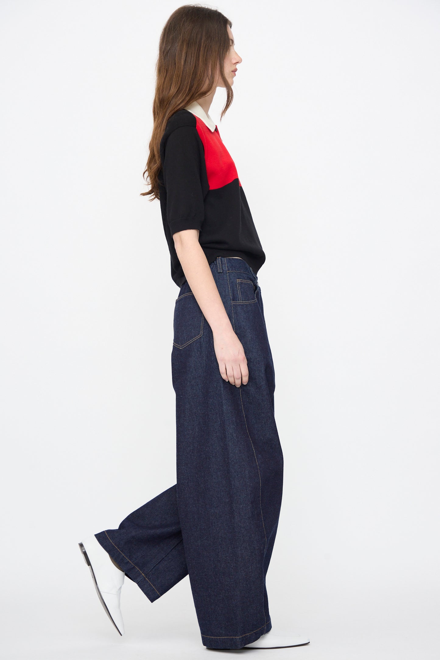 A person stands in profile wearing Cordera Curved Baggy Jeans in Denim, a black and red top, and white pointed shoes against a plain white background.