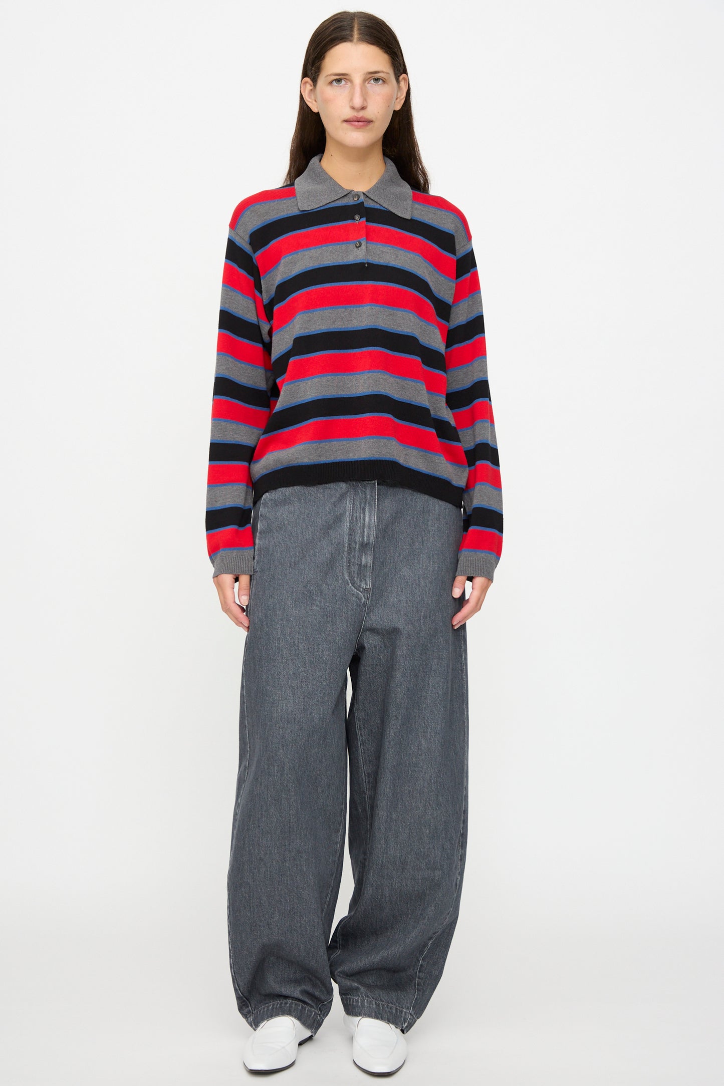 A person standing against a white background wearing a red, navy, and gray striped sweater, Curved Pants in Washed Denim by Cordera styled in an oversized wide-leg fit, and white shoes.