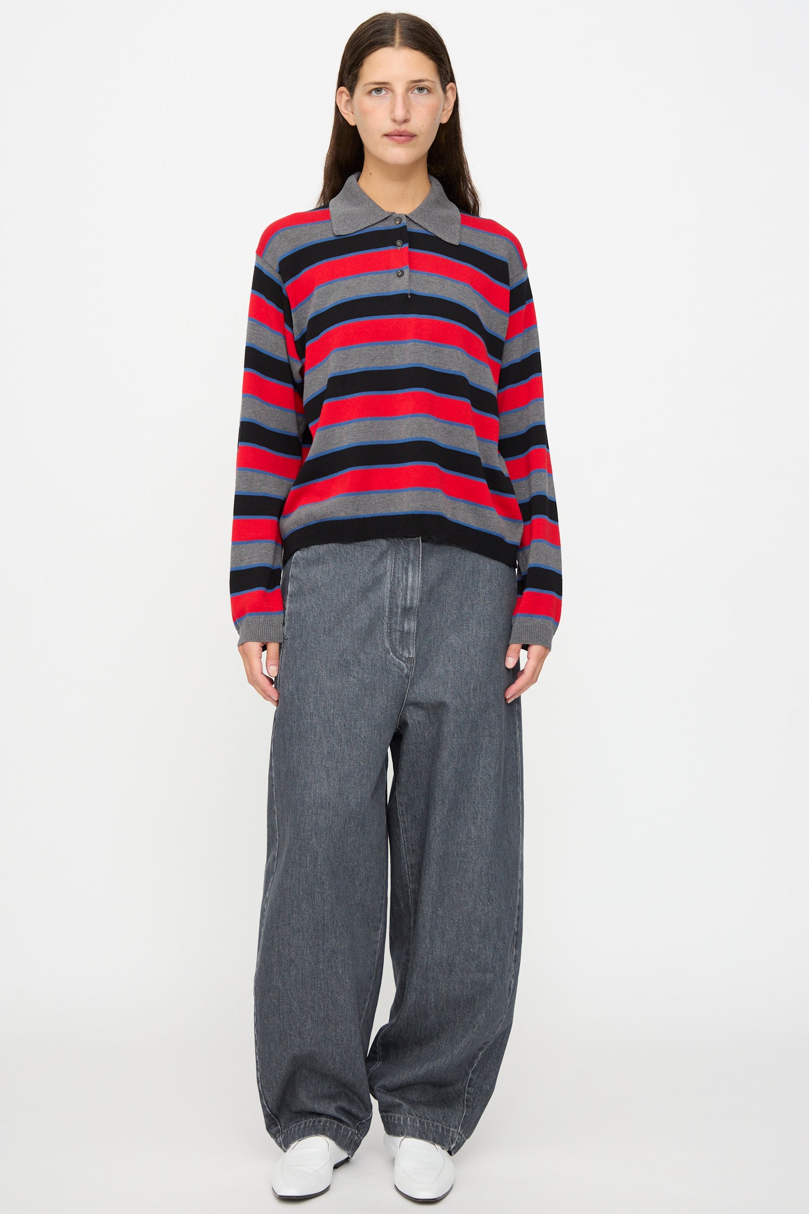A person standing against a white background wearing a red, navy, and gray striped sweater, Curved Pants in Washed Denim by Cordera styled in an oversized wide-leg fit, and white shoes.