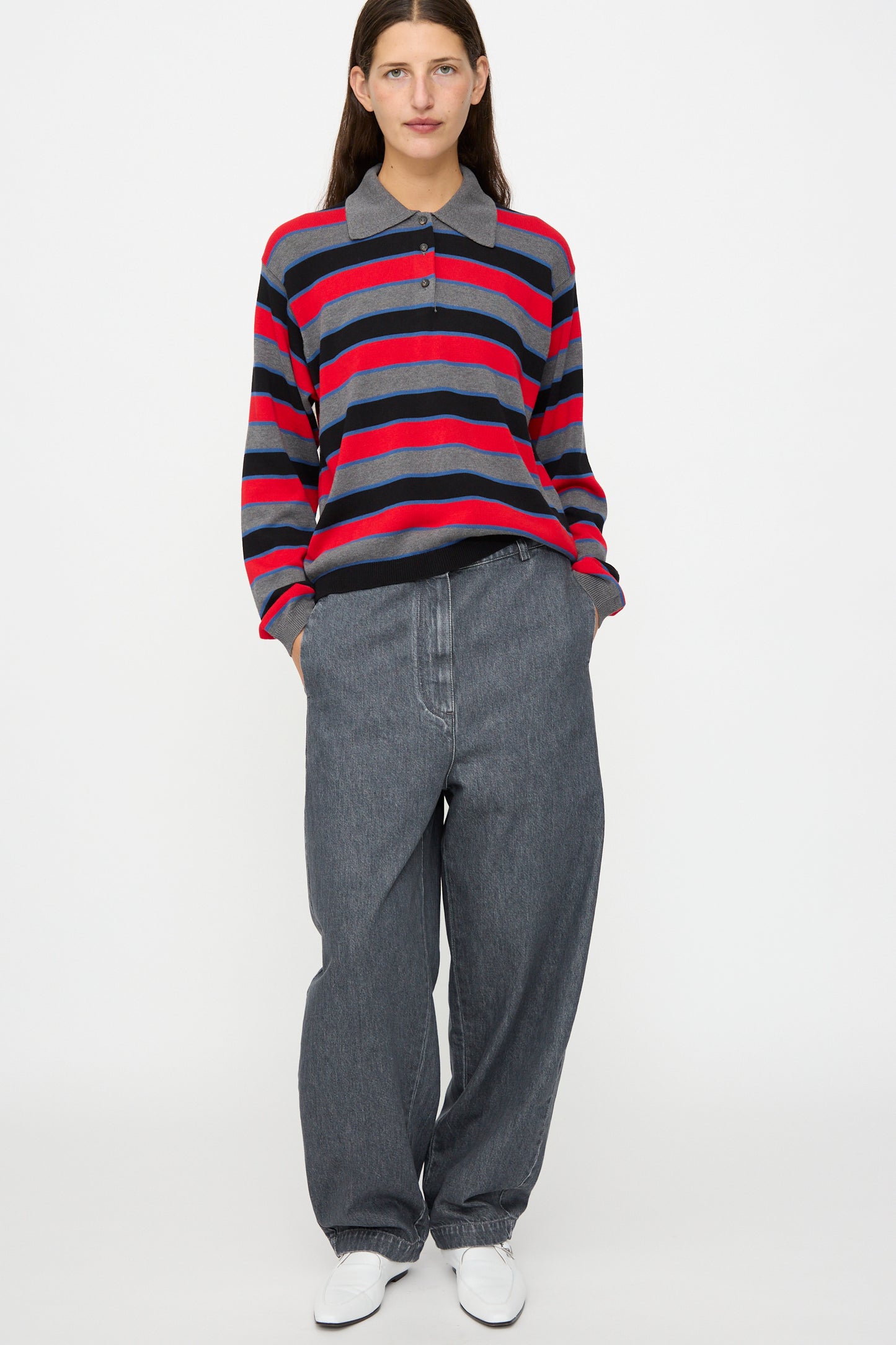 A person stands facing forward against a plain white background, wearing Cordera’s Curved Pants in Washed Denim, a red and gray striped long-sleeve polo shirt, and white shoes, with their hands in their pockets.