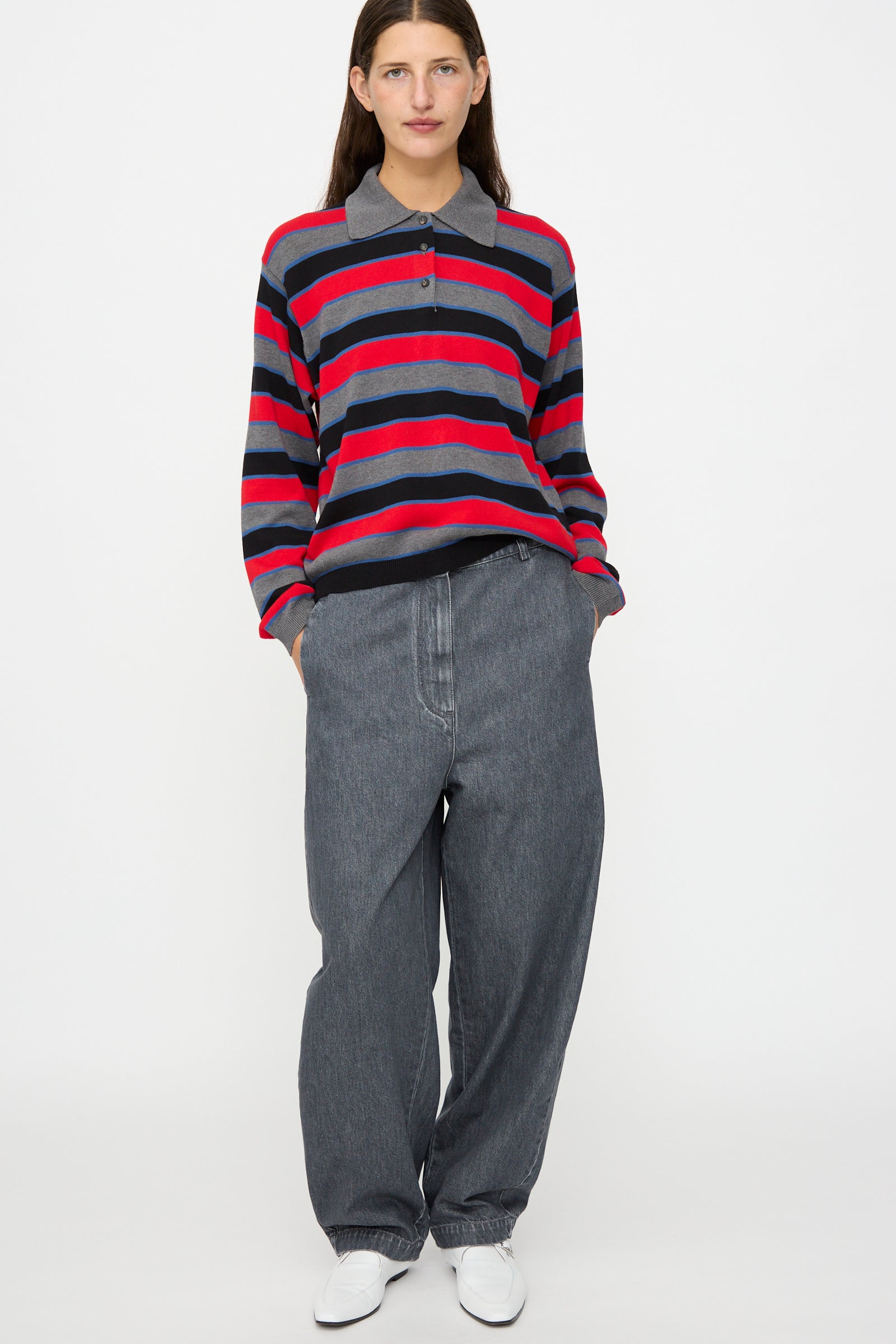 A person stands facing forward against a plain white background, wearing Cordera’s Curved Pants in Washed Denim, a red and gray striped long-sleeve polo shirt, and white shoes, with their hands in their pockets.