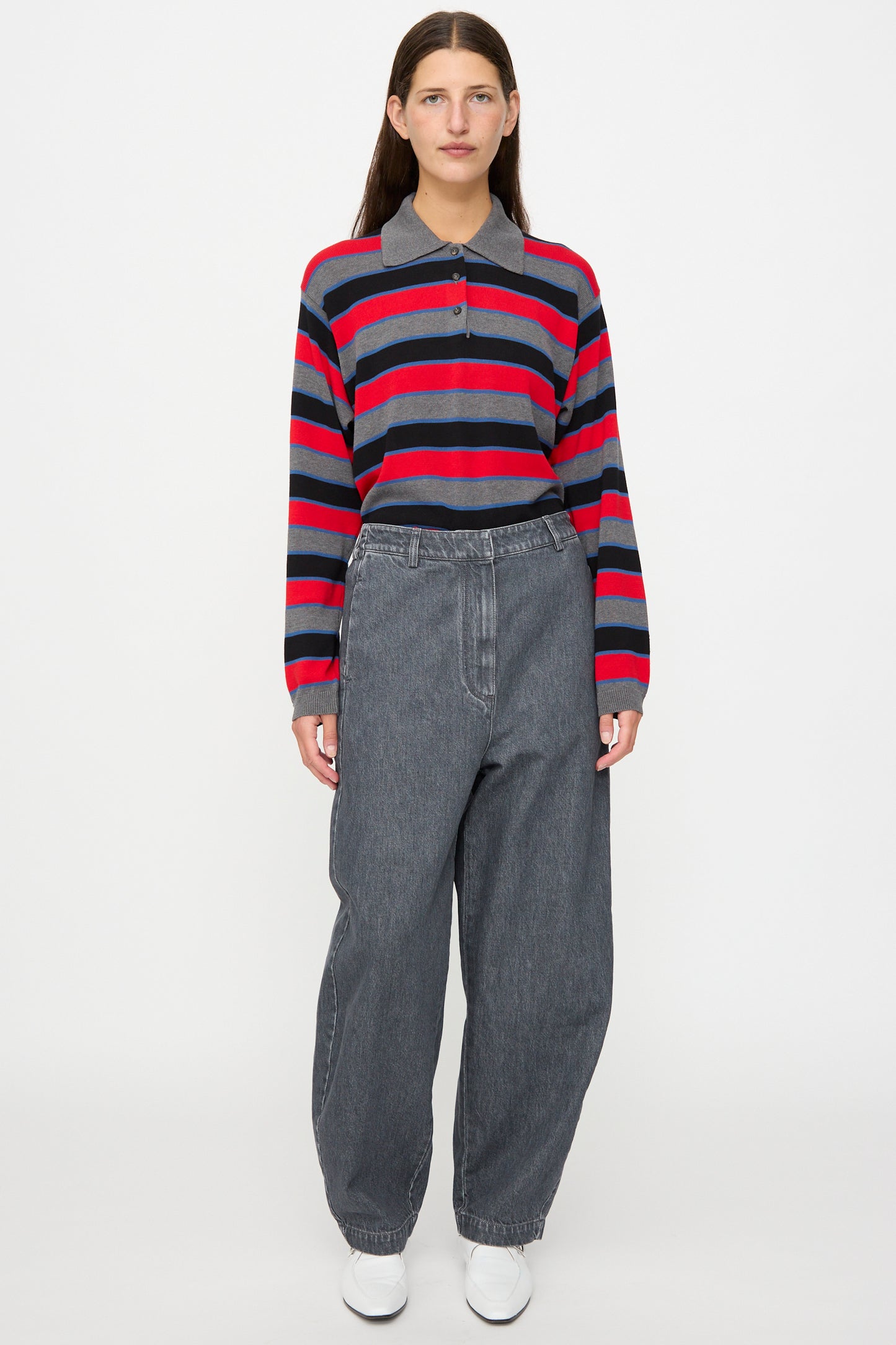 A person stands against a plain background, wearing Cordera’s Curved Pants in Washed Denim paired with a red and navy striped long-sleeve shirt and white shoes.