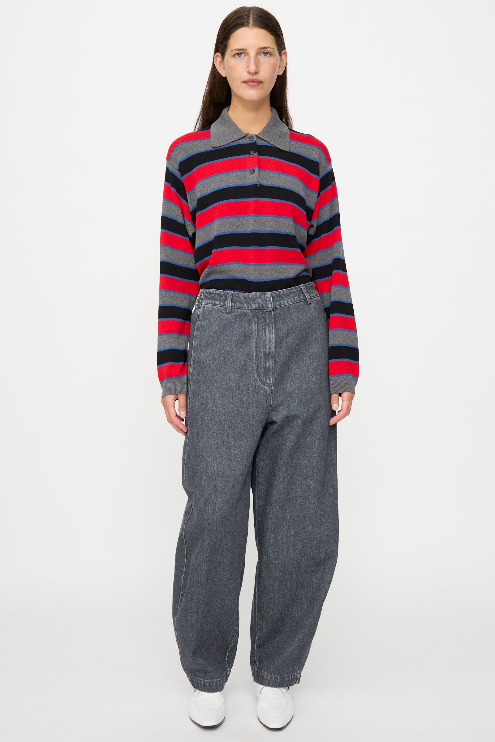 A person stands against a plain background, wearing Cordera’s Curved Pants in Washed Denim paired with a red and navy striped long-sleeve shirt and white shoes.