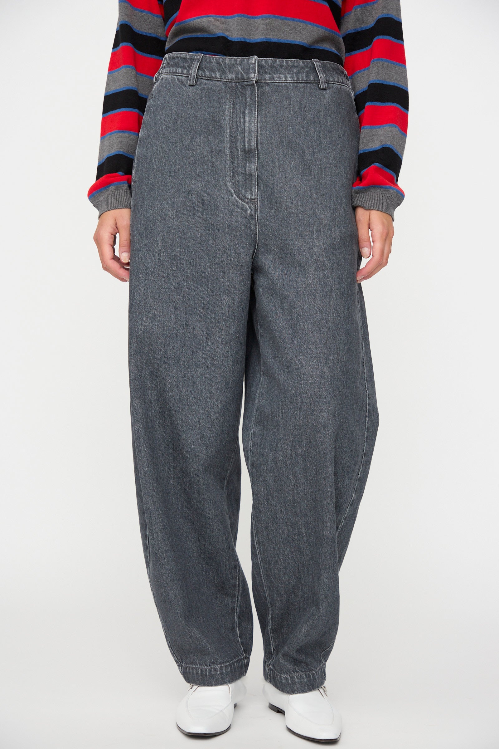 A person is wearing the Cordera Curved Pants in Washed Denim—high-waisted, wide-leg gray jeans—paired with a striped long-sleeve shirt in red, black, and gray, and white shoes, standing against a plain background.