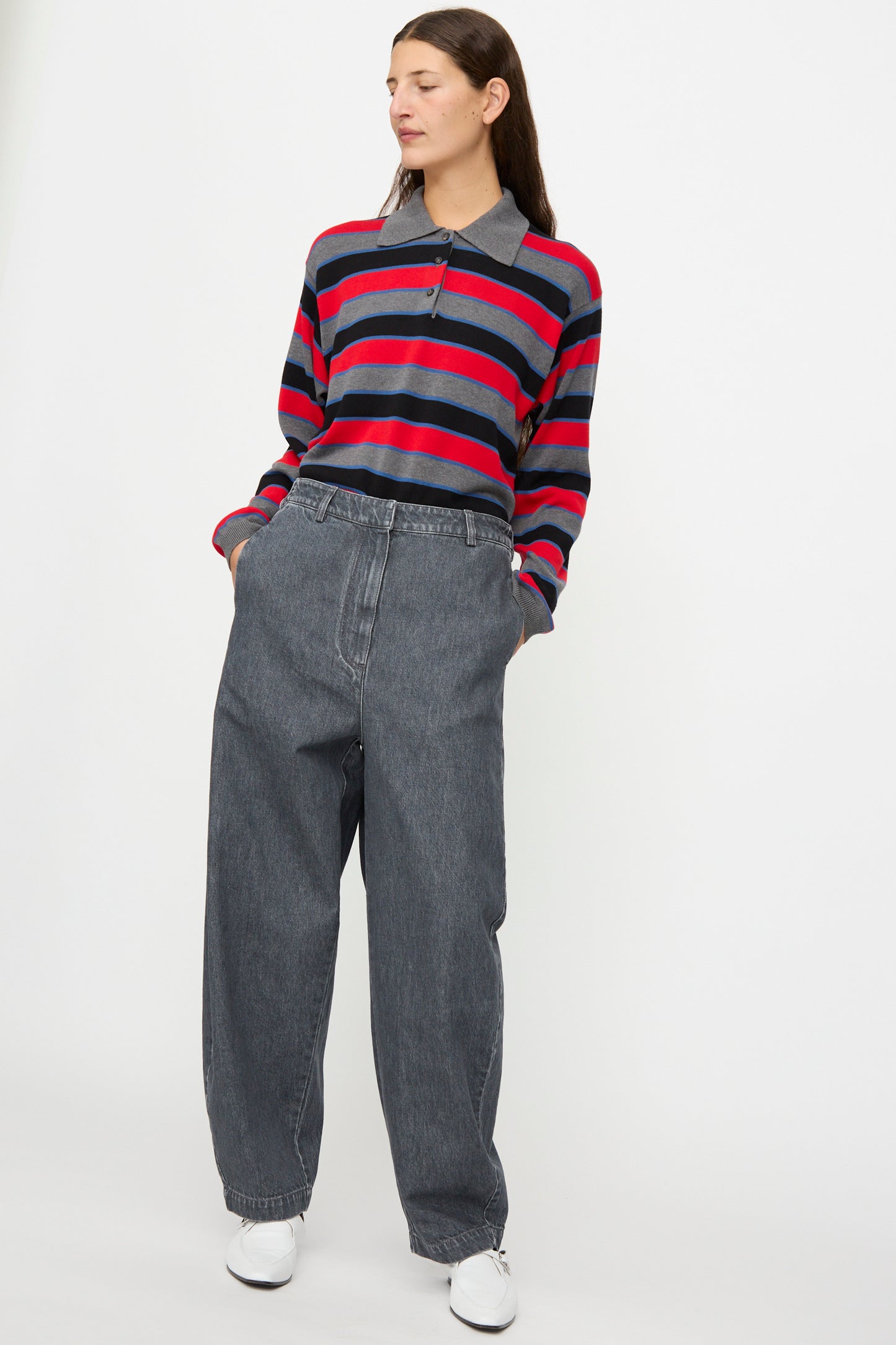 A person stands with hands in pockets against a plain white background, wearing a red and navy striped long-sleeve shirt, Cordera Curved Pants in Washed Denim, and white sneakers.