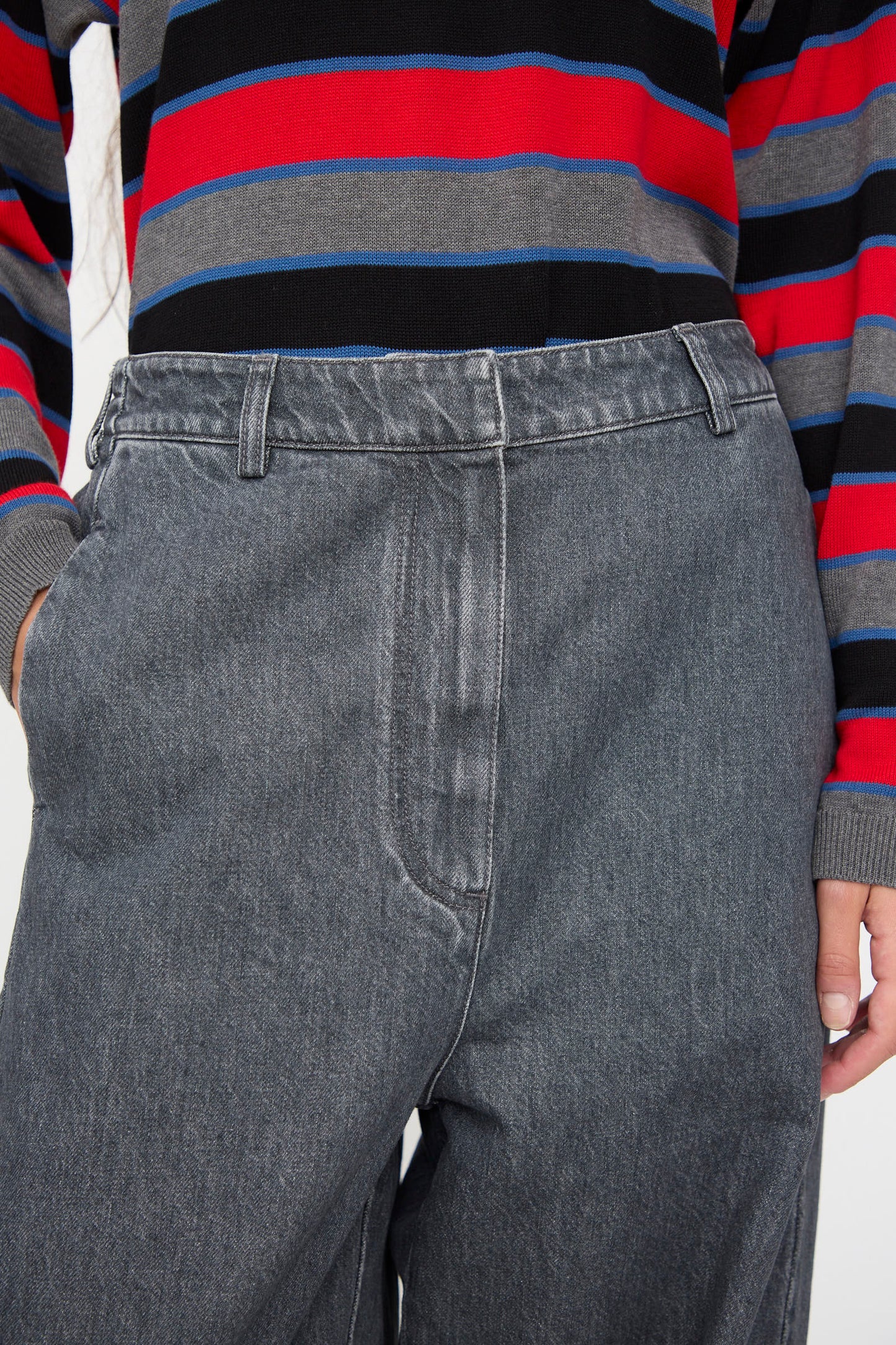 A person wearing Cordera’s Curved Pants in Washed Denim—high-waisted, wide leg gray jeans—paired with a long-sleeve shirt featuring red, blue, gray, and black horizontal stripes, hand casually placed in their pocket.