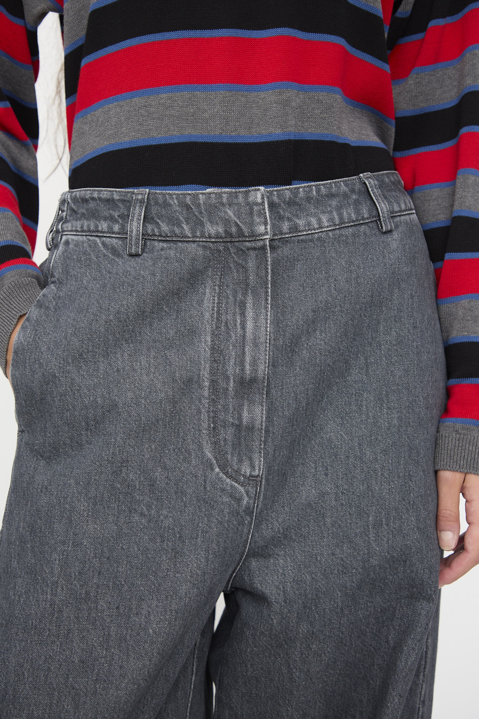 A person wearing Cordera’s Curved Pants in Washed Denim—high-waisted, wide leg gray jeans—paired with a long-sleeve shirt featuring red, blue, gray, and black horizontal stripes, hand casually placed in their pocket.