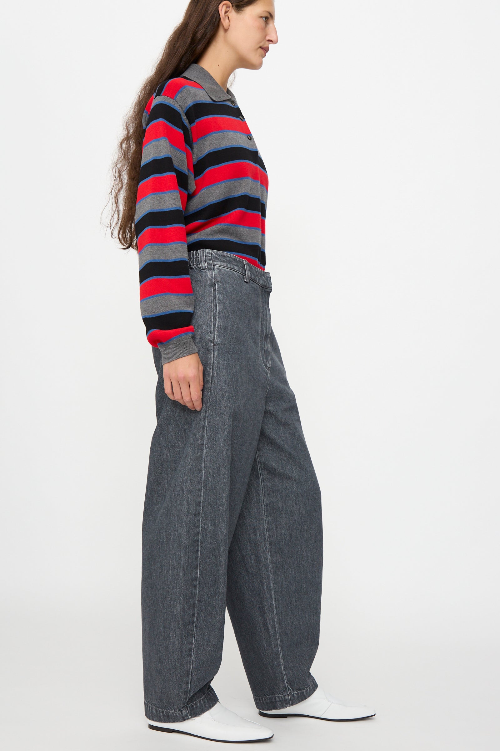 A person stands sideways against a plain white background, wearing Cordera’s Curved Pants in Washed Denim paired with a gray, red, and navy striped sweater and white shoes.