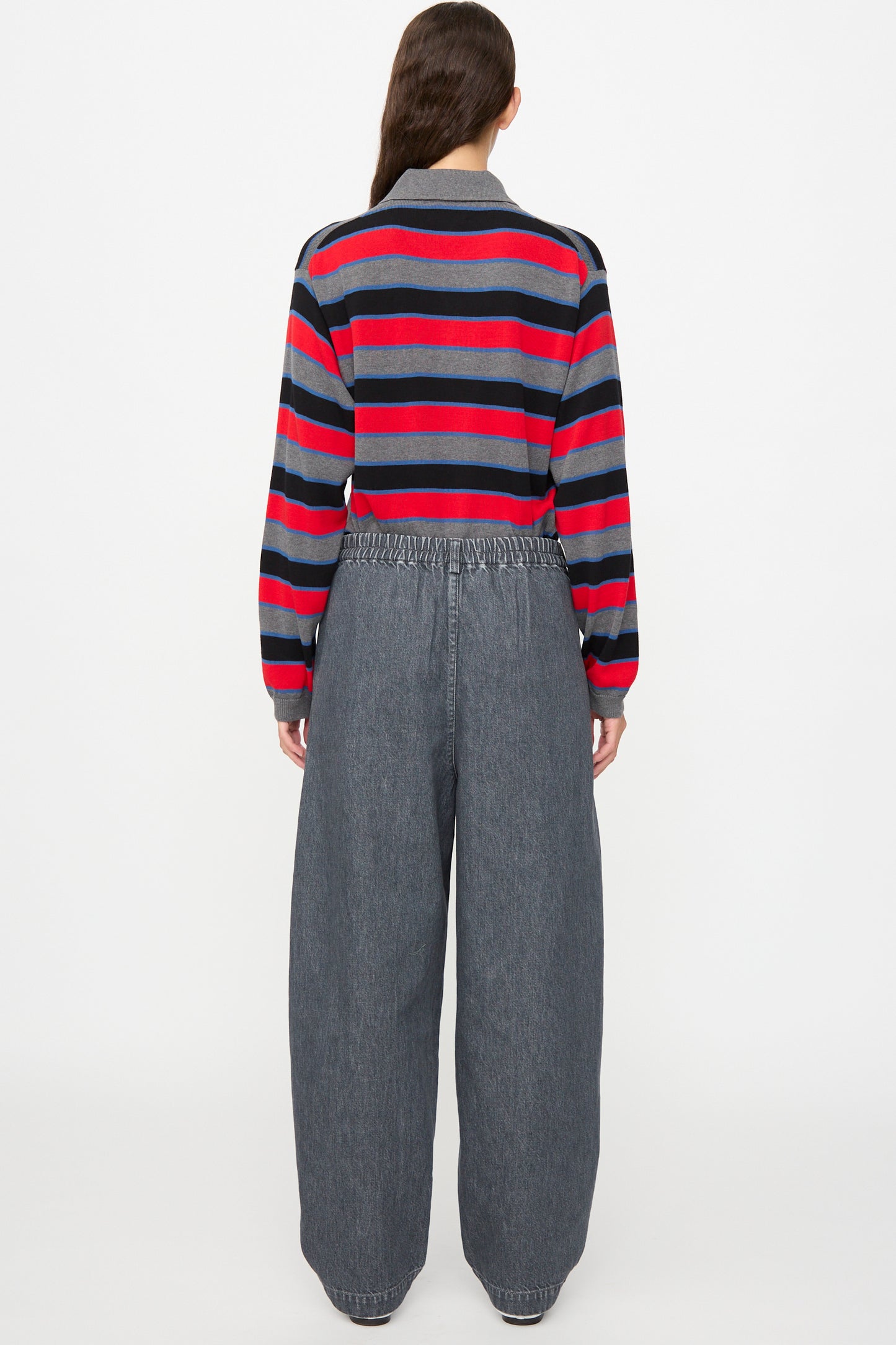 A person stands with their back to the camera, wearing Cordera’s Curved Pants in Washed Denim and a gray and red striped long-sleeve shirt, set against a plain white background.