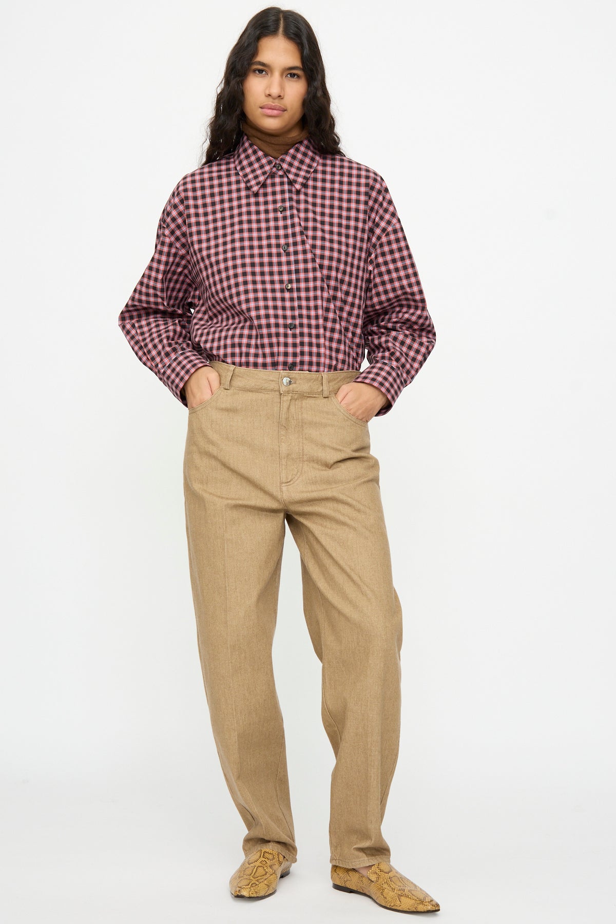 A person wearing a long-sleeved red and black plaid button-down shirt tucked into Cordera Denim Carrot Pants in Beige with their hands in their pockets.