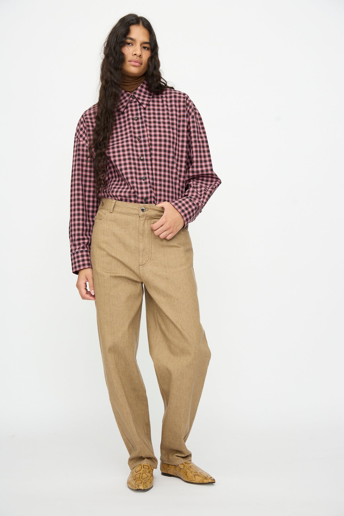 A person wearing a long-sleeved red and black checkered button-down shirt tucked into loose-fitting, tapered Denim Carrot Pants in Beige by Cordera.