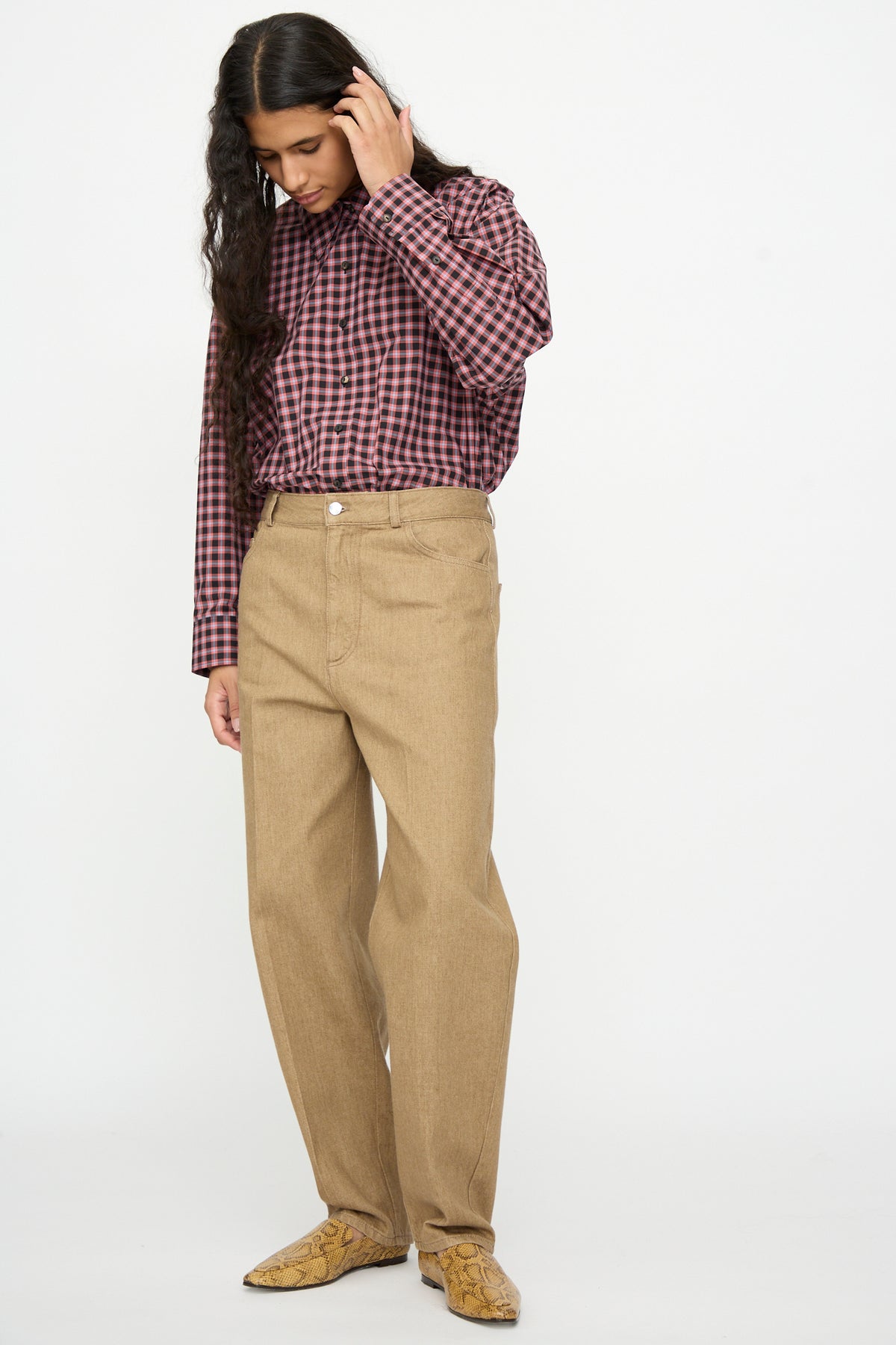 A person wearing a pink and black plaid button-down shirt and beige Denim Carrot Pants in a high-waisted tapered leg fit with snakeskin-print shoes.