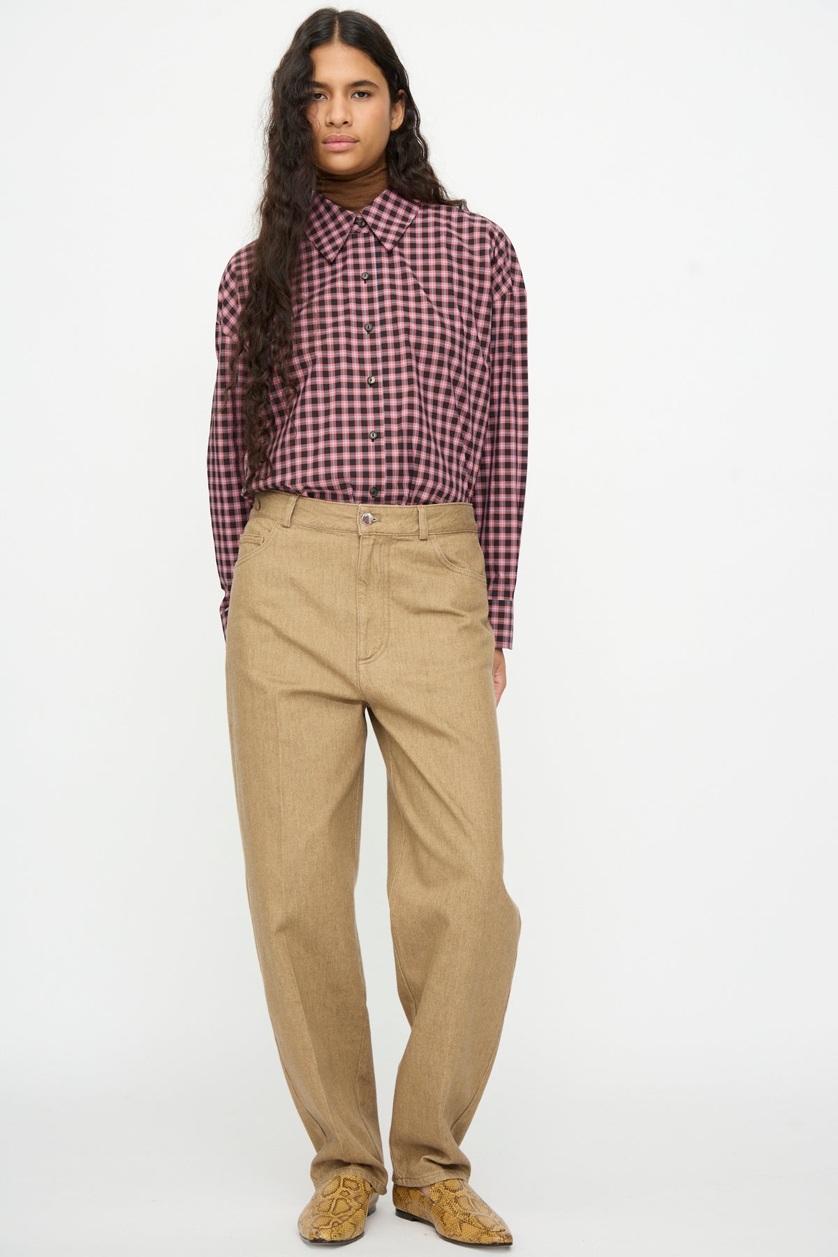 A person wearing a pink and black plaid button-down shirt tucked into beige Cordera Denim Carrot Pants with a high-waisted, tapered-leg fit.