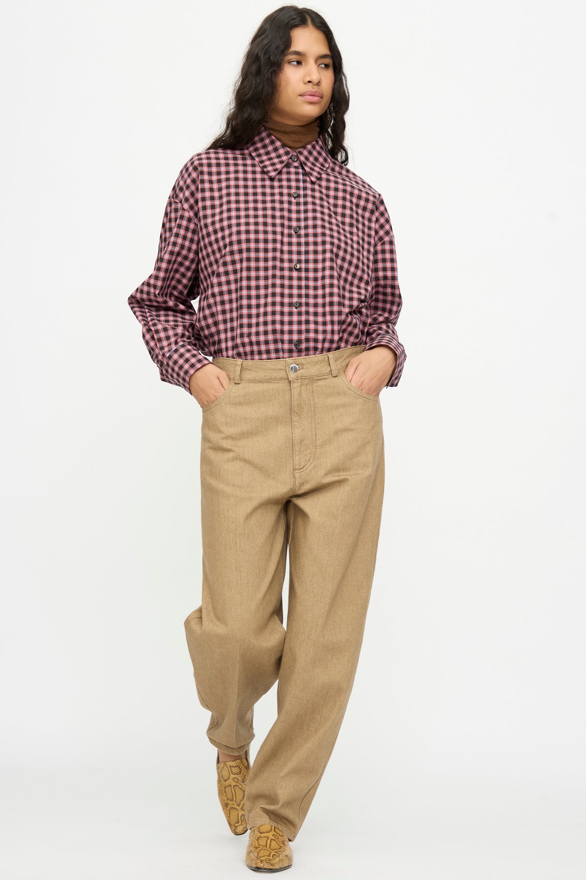 A person wearing a pink and black plaid button-down shirt tucked into beige Denim Carrot Pants by Cordera with their hands in their pockets.