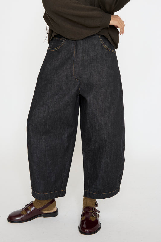 A person models the Cordera Denim Curved Pant in Black, featuring a wide-leg and 5-pocket design, made from organic cotton. They pair it with a dark top, brown socks, and burgundy shoes against a plain background.