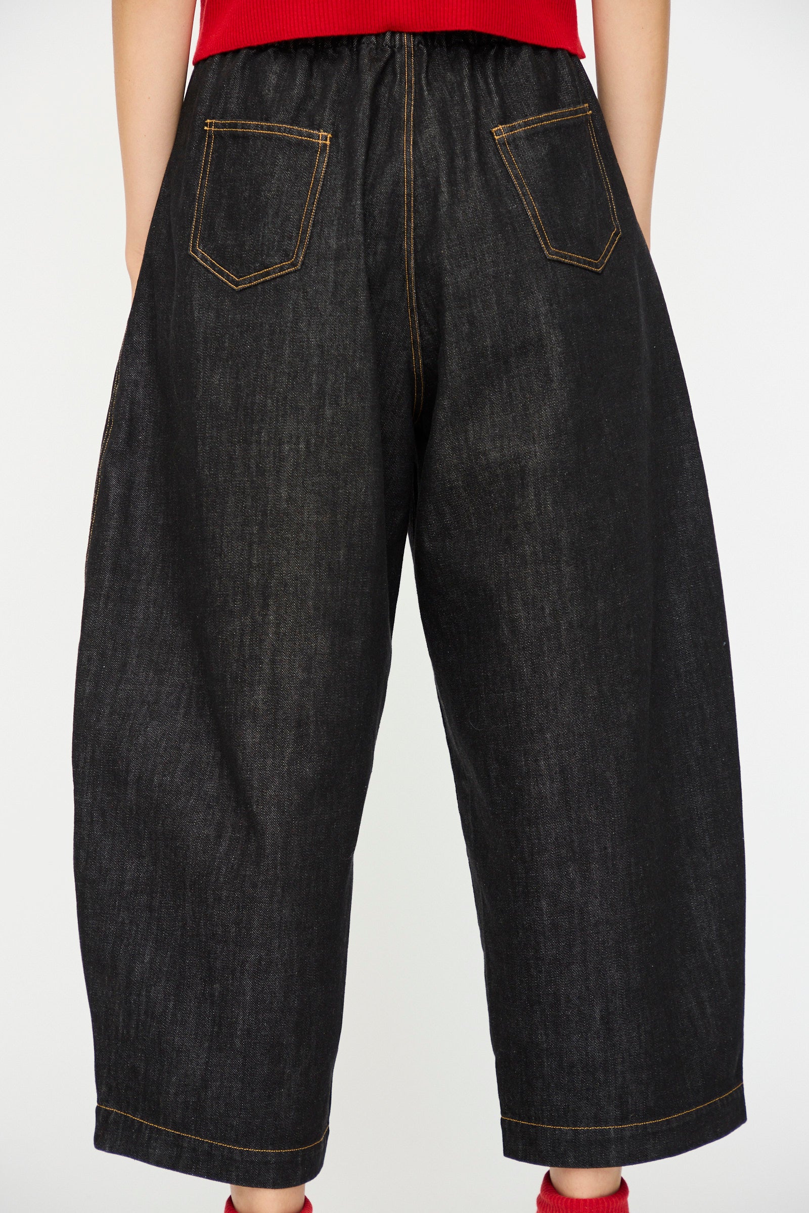 A person wears Cordera's Denim Curved Pant in Black—loose-fitting, dark organic cotton denim jeans with an elastic waistband and yellow stitching—standing against a plain white background.