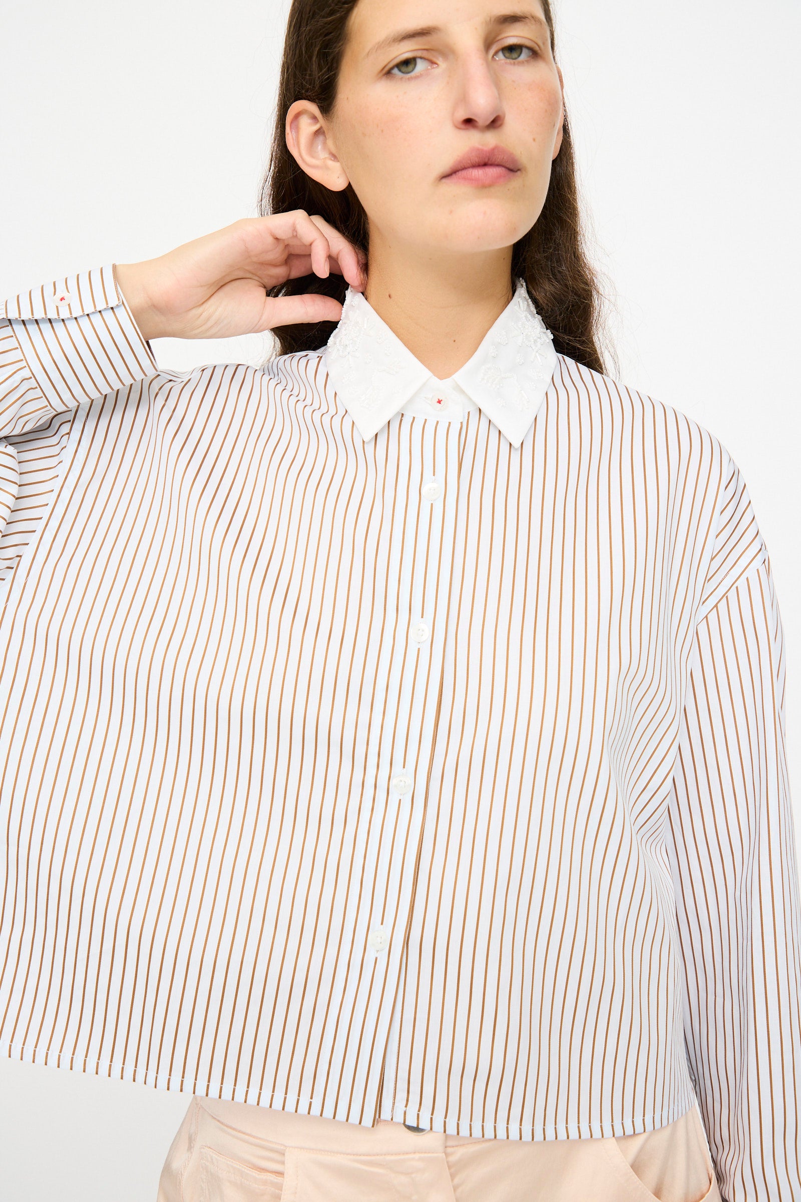 A person wearing Cordera's Embellished Shirt in Striped, featuring long sleeves, vertical brown stripes, and a white collar, poses with one hand touching their neck.