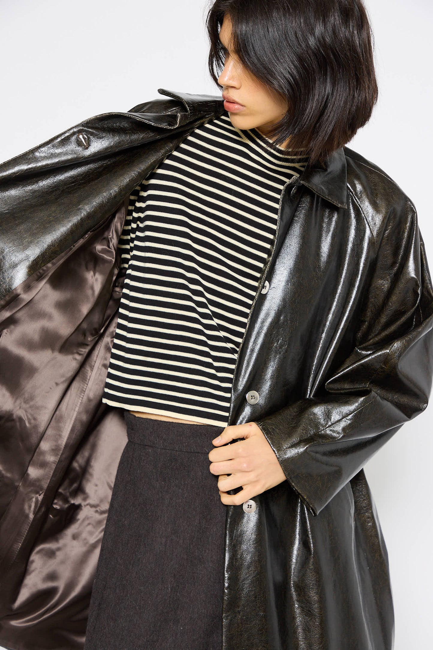 A person wearing a Cordera Faux Scratched Leather Coat in Black over a black and white striped shirt and dark pants looks off to the side.