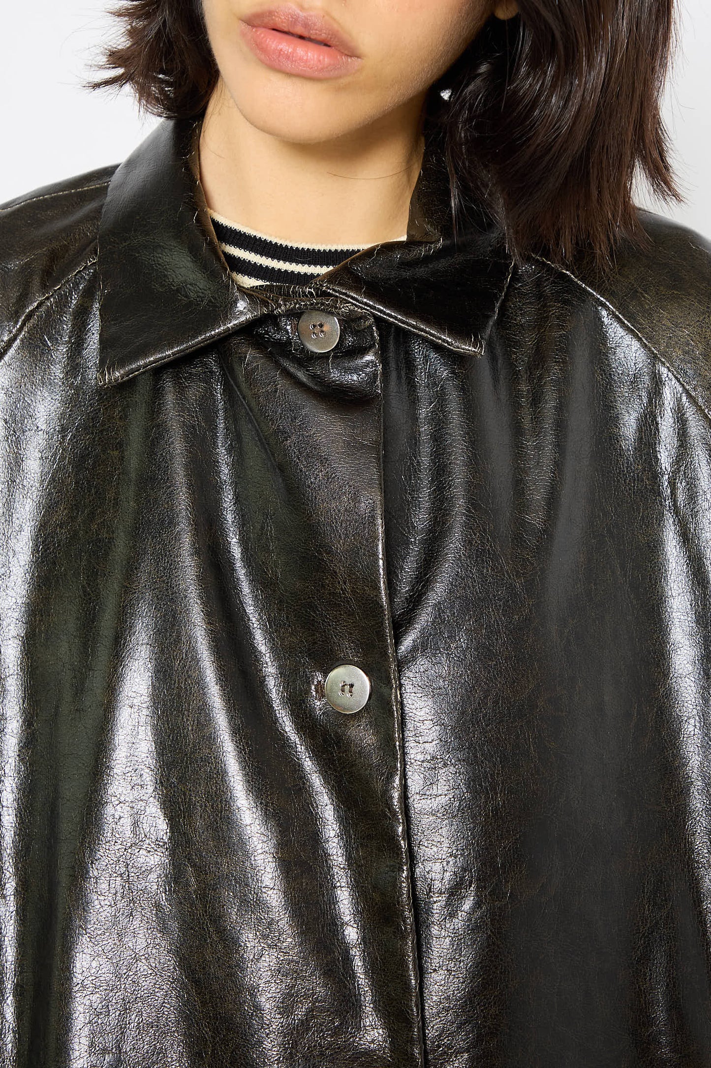 A close-up image features a person wearing a Cordera Faux Scratched Leather Coat in Black, showcasing its shiny texture. The person has short dark hair and is donning a black and white striped shirt underneath.