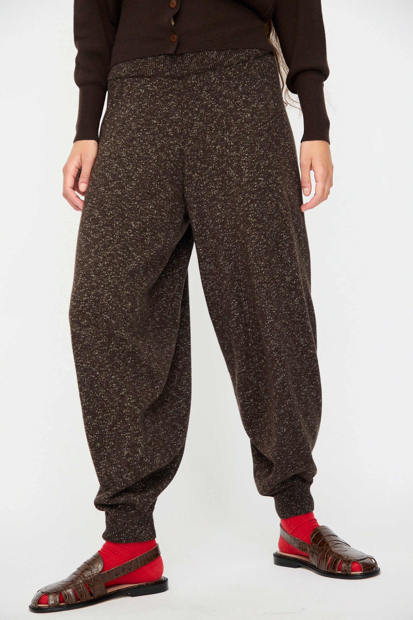 A person wears Cordera's Flecked Cotton Pant in Brown, paired with a dark top, red socks, and brown sandals, standing against a plain white background.