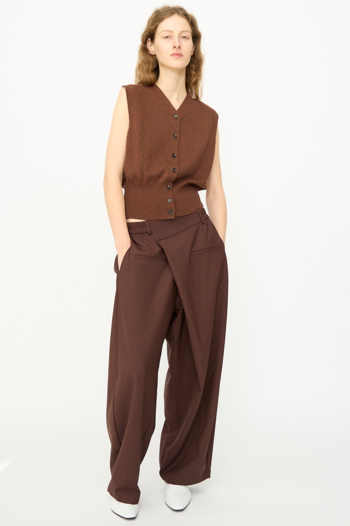 A person stands against a plain background wearing Cordera’s Flecked Cotton Waistcoat in Russet, paired with loose brown draped pants and white shoes.