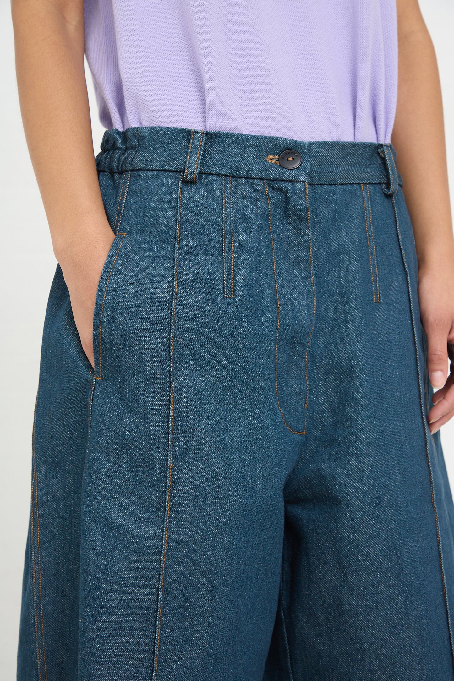 Close-up of a person wearing a lilac top and Cordera's Frontal Seam Curved Pant in Denim jeans with a button and stitching details.