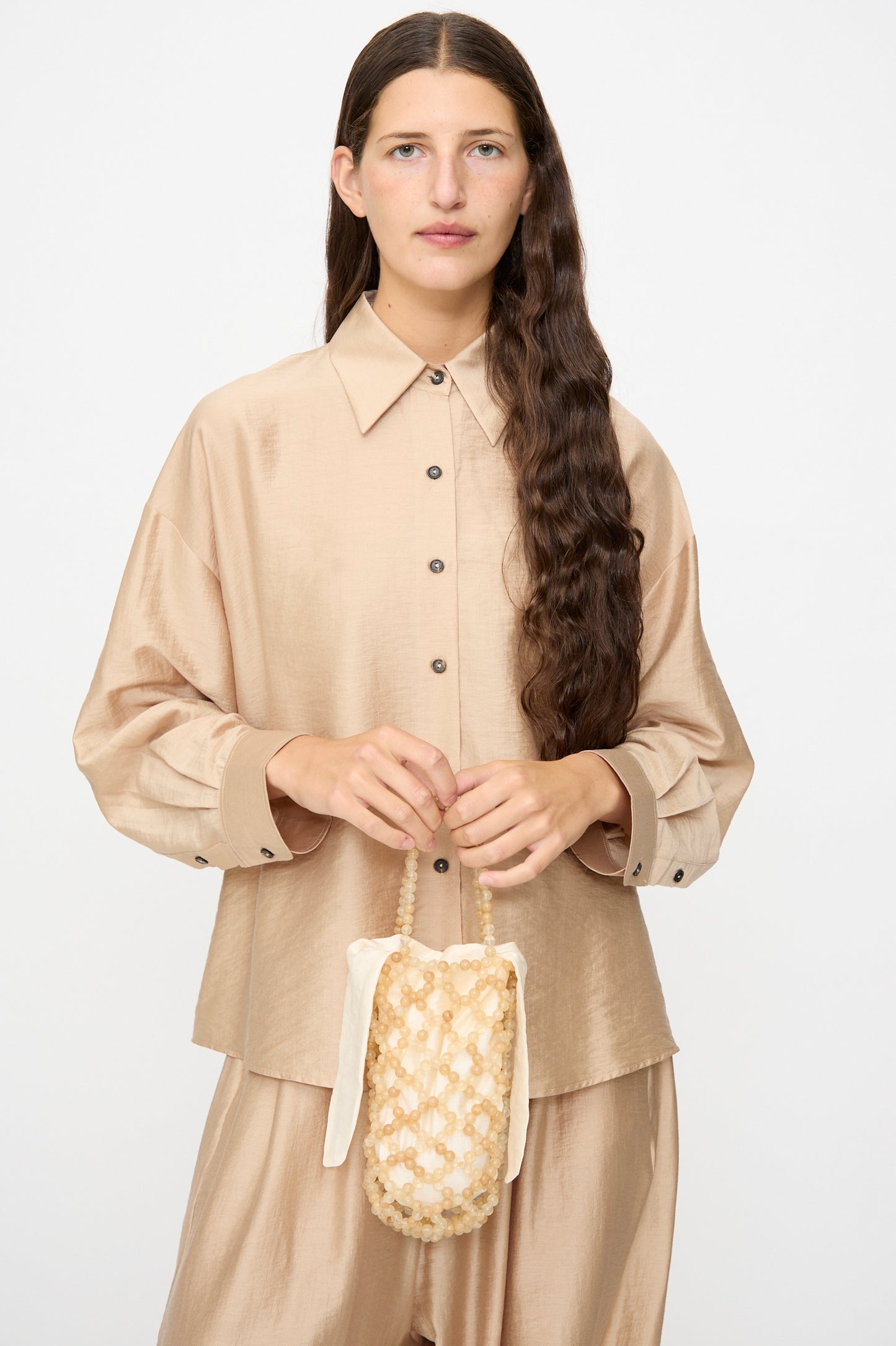 A woman with long brown hair wears a beige button-up shirt and pants, holding the Cordera Handmade Beaded Bag in Jasper Miel—crafted in Guatemala with cream and beige woven detailing and a handle—while standing in front of a plain light background.