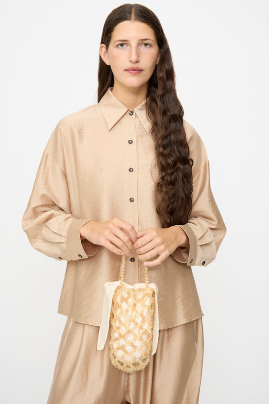 A woman with long brown hair wears a beige button-up shirt and pants, holding the Cordera Handmade Beaded Bag in Jasper Miel—crafted in Guatemala with cream and beige woven detailing and a handle—while standing in front of a plain light background.